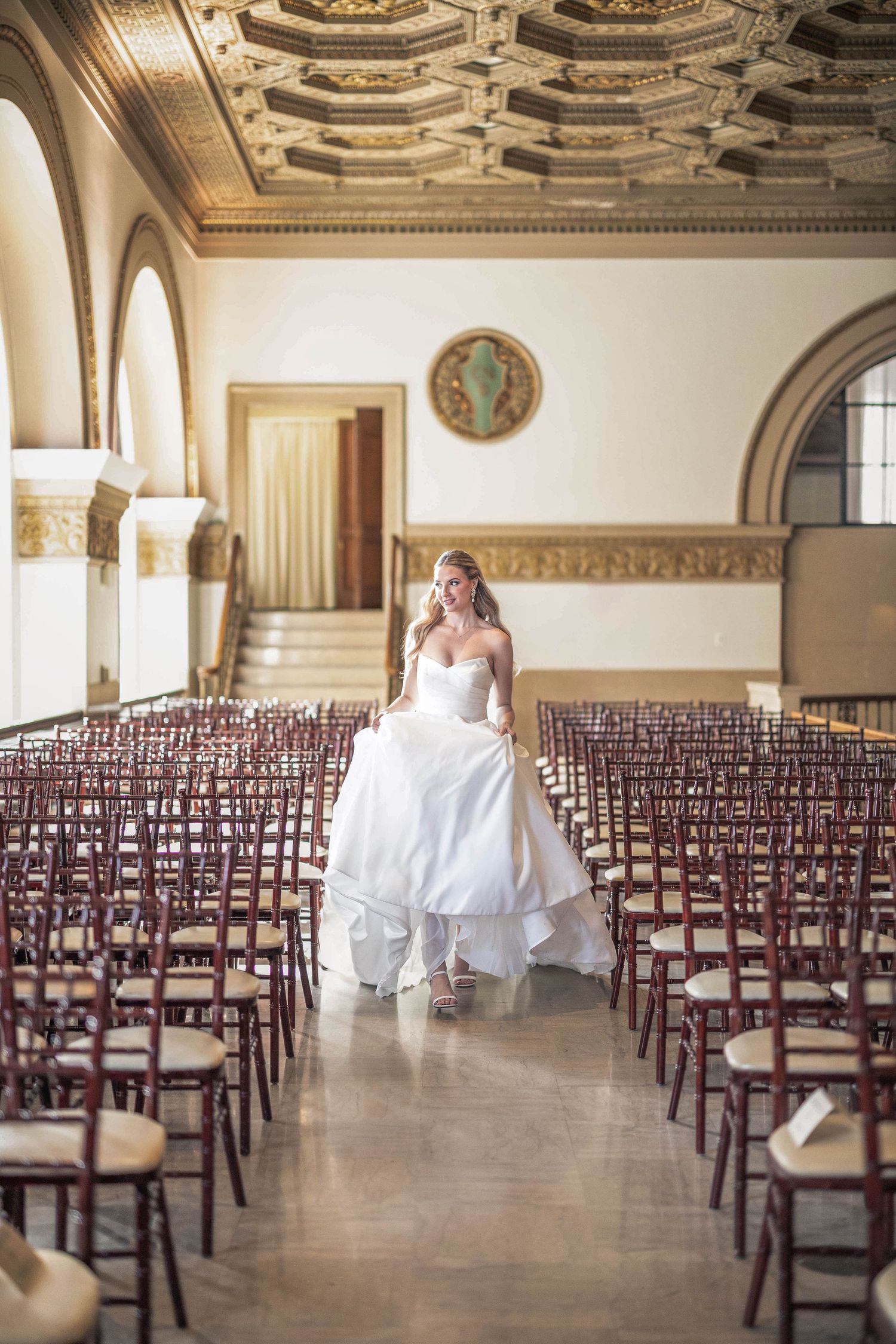 Bride in white strapless ball gown walks down aisle of ornate wedding venue with rows of wooden chairs.