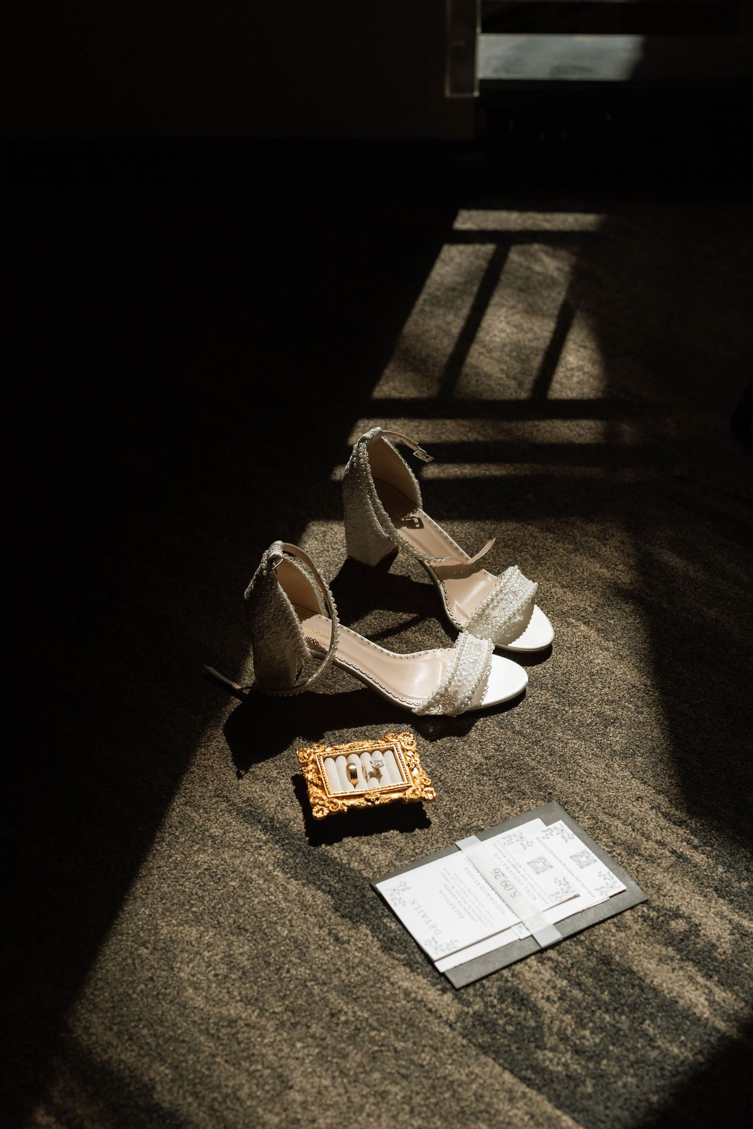 Bridal heels, rings in gold frame, and wedding vows lit by dramatic sunlight on a carpeted floor.