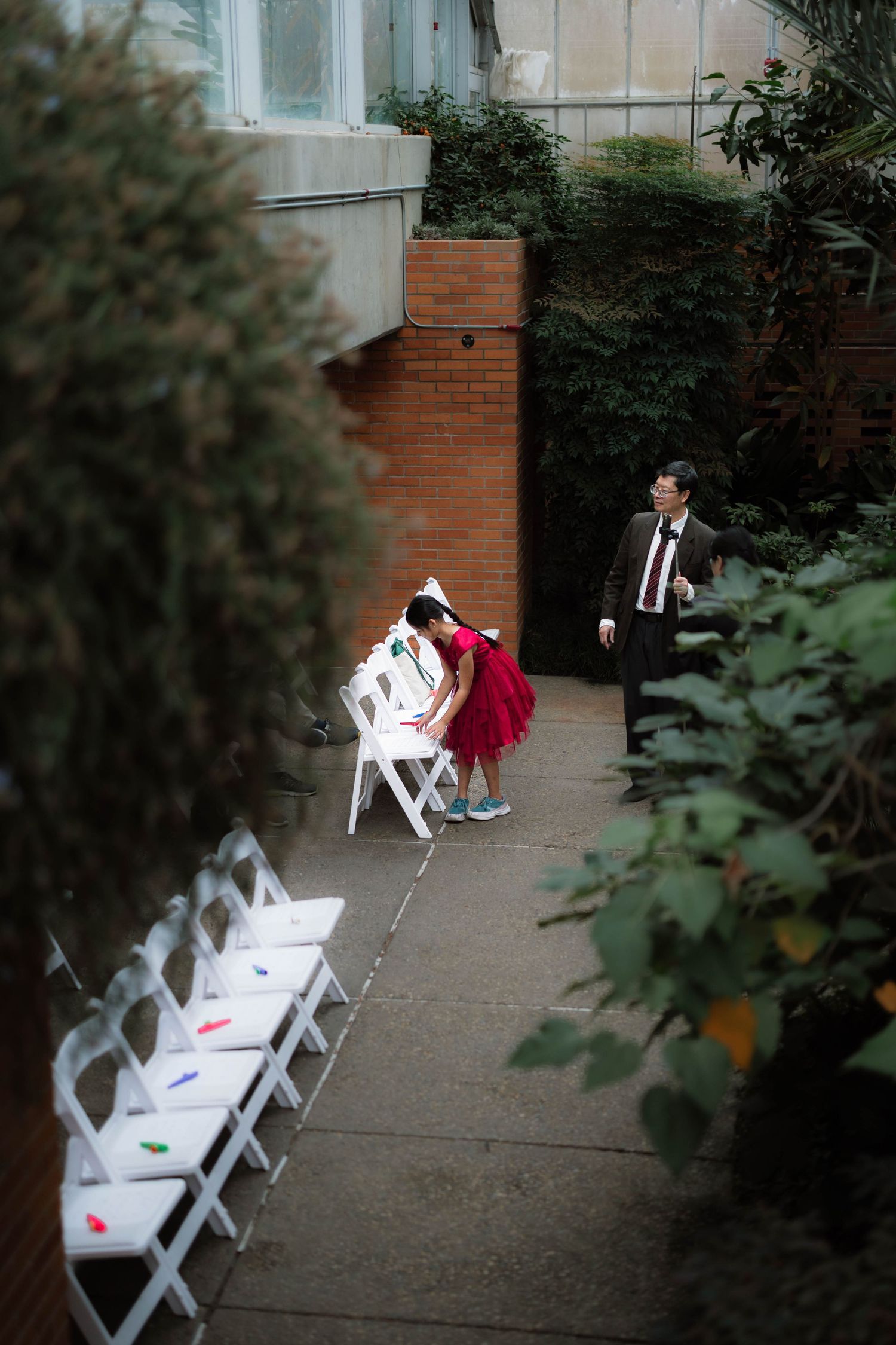 Child in red dress bends over white chairs at outdoor wedding venue as man in suit looks on.