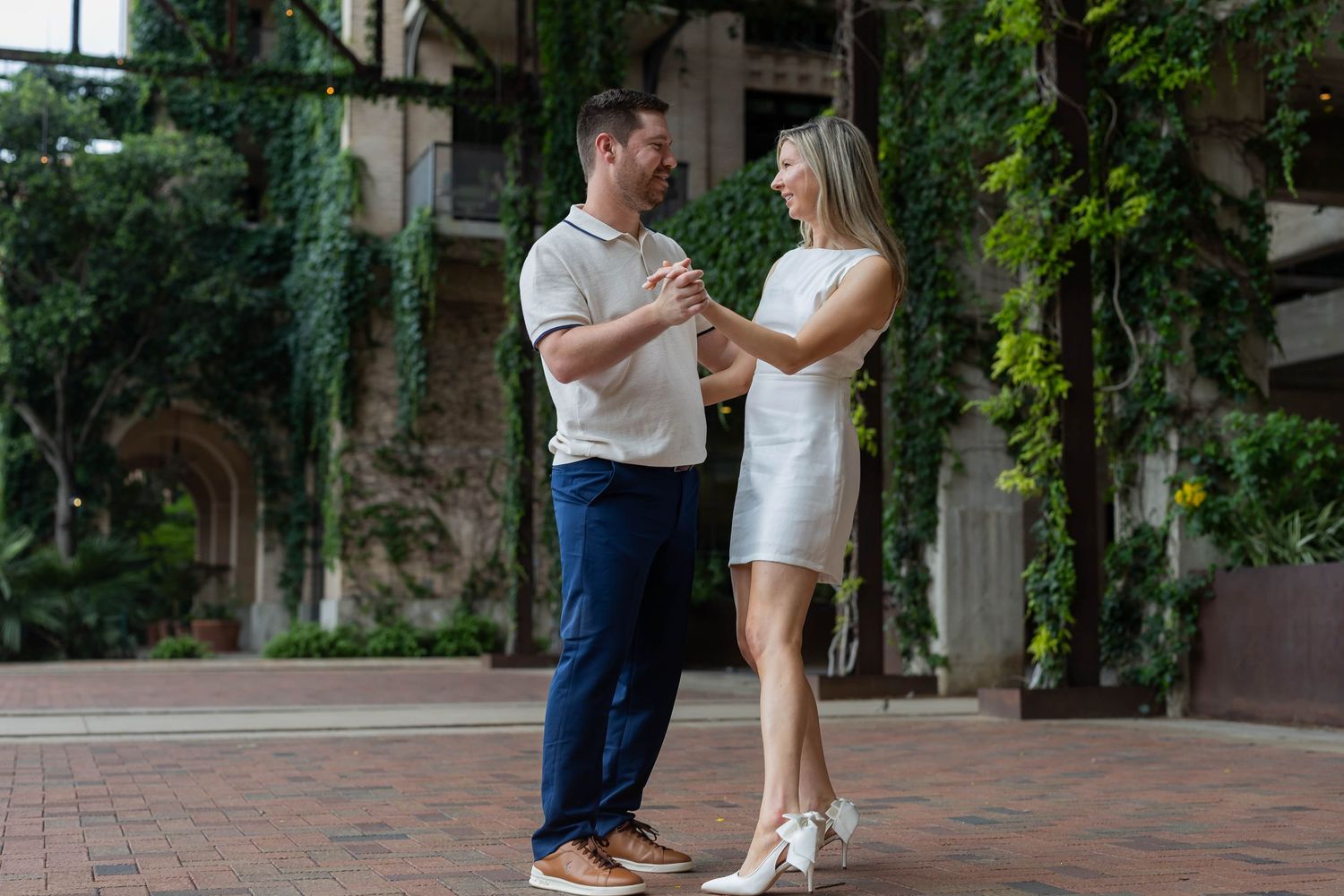 Couple holding hands and dancing outdoors near ivy-covered building on brick patio in romantic engagement photo.