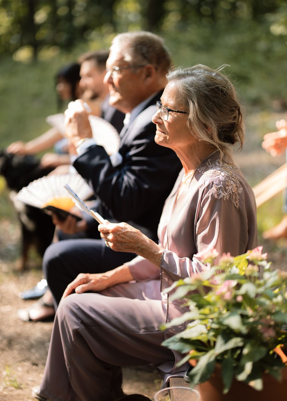 Elderly woman smiling and holding a program at an outdoor wedding ceremony, seated among other guests in sunlight.
