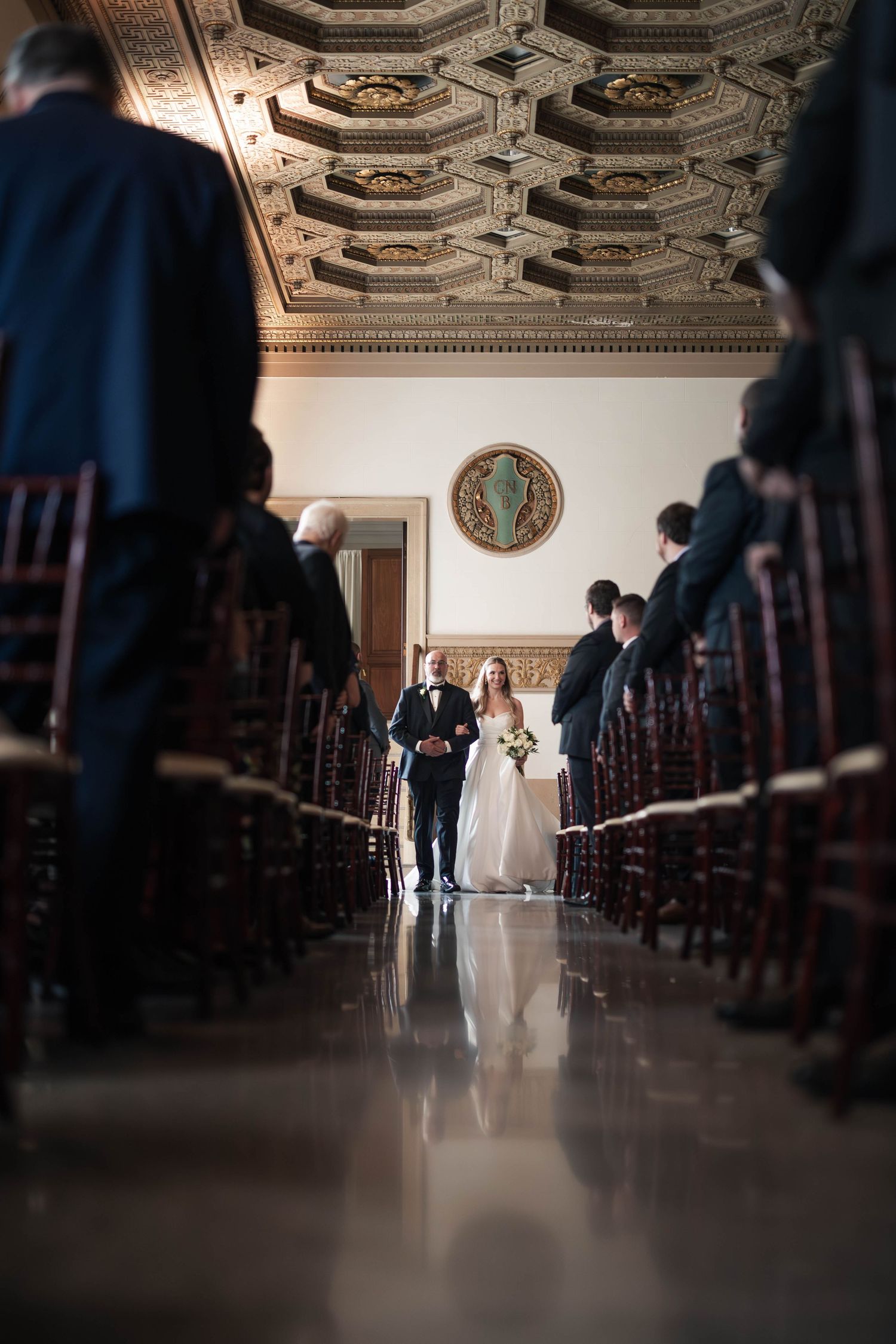 Bride walks down aisle with escort in grand ornate ceremony hall with coffered ceiling and wooden chairs.