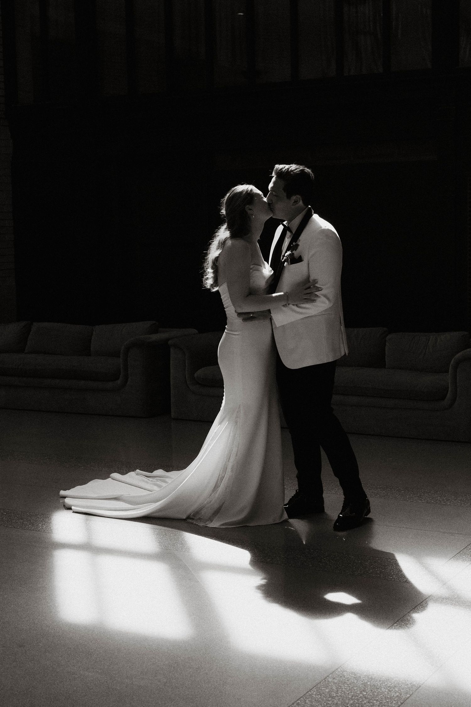 Bride and groom share first dance kiss in dramatic black and white light.