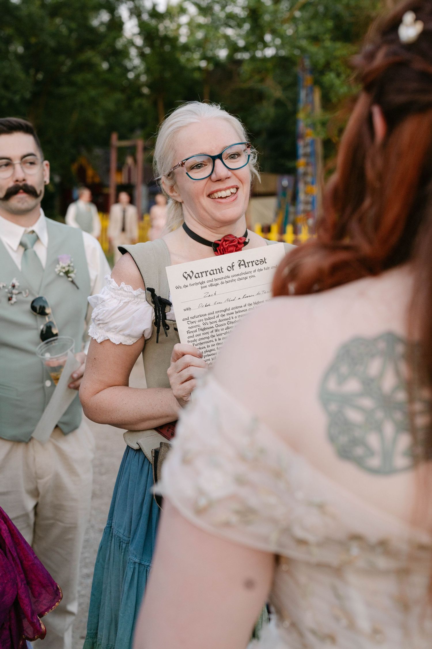 Smiling woman with glasses holds a Warrant of Arrest prop at a themed outdoor wedding ceremony.