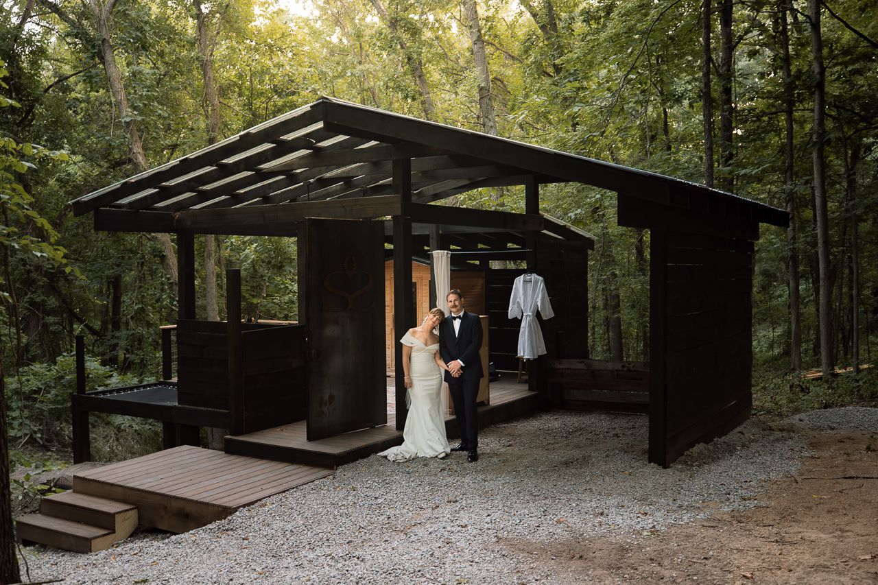 Bride and groom embrace outside a modern dark wood cabin surrounded by lush forest trees on their wedding day.