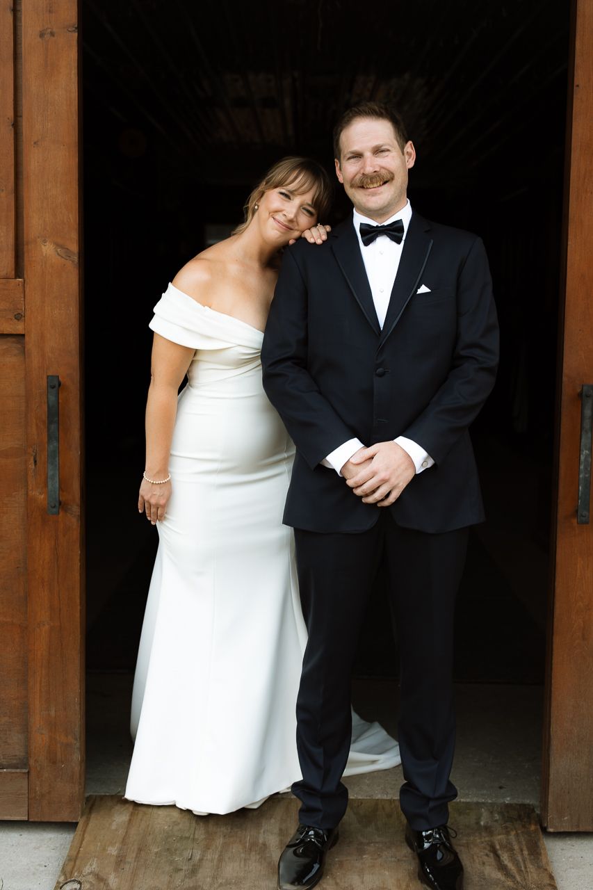 Bride in off-shoulder white gown and groom in black tuxedo posing together at wooden barn doors on wedding day.