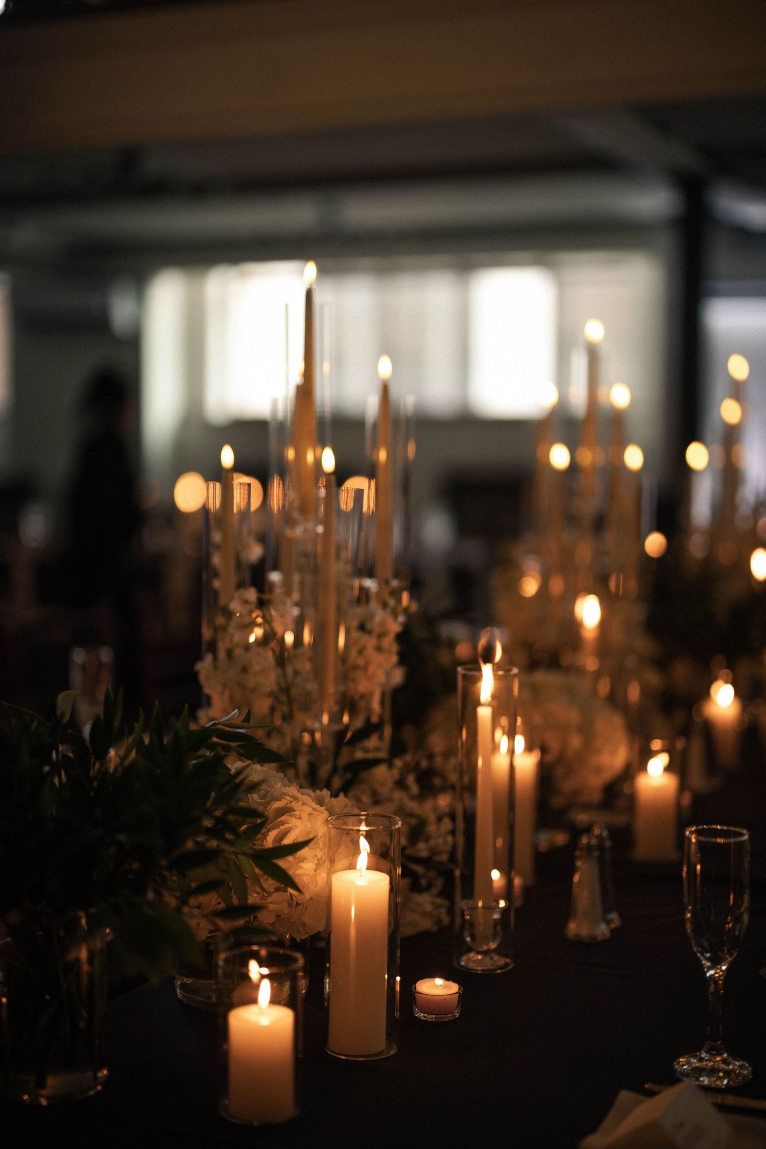 Romantic wedding reception table adorned with glowing white pillar candles and floral centerpieces in dim lighting.