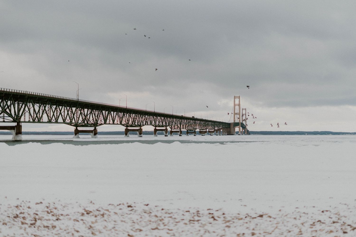 A long bridge spans across a snow-covered winter landscape under cloudy skies.