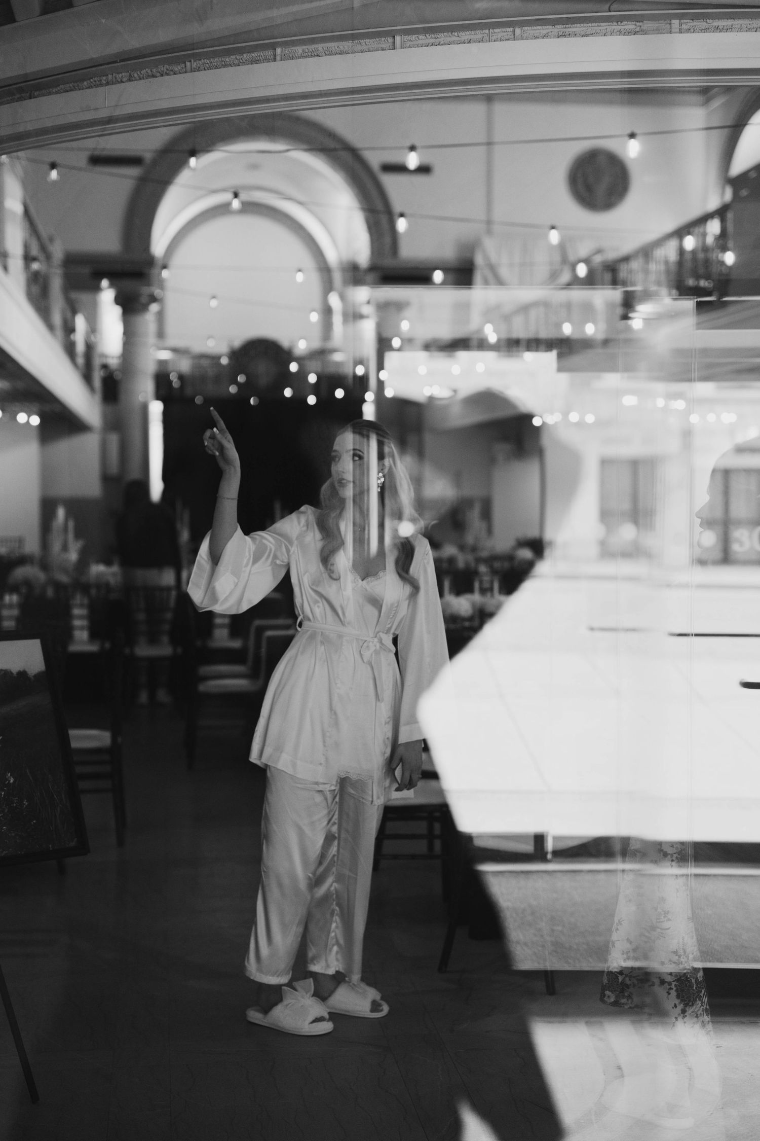 Black and white photo of a woman in pajamas and slippers standing inside an elegant restaurant with string lights.