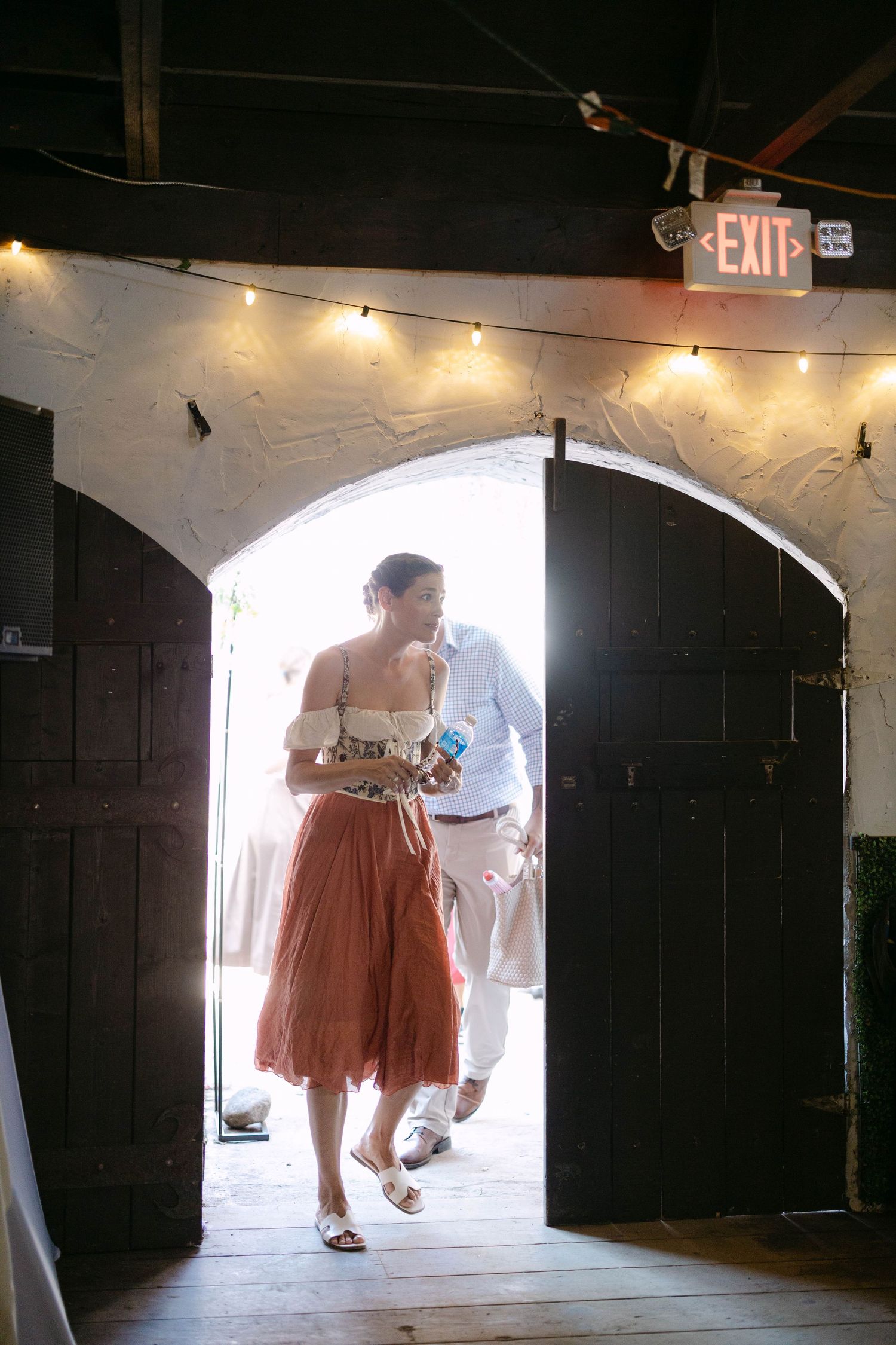 Couple entering a rustic wedding venue through arched wooden doors with warm string lights and glowing backlight.