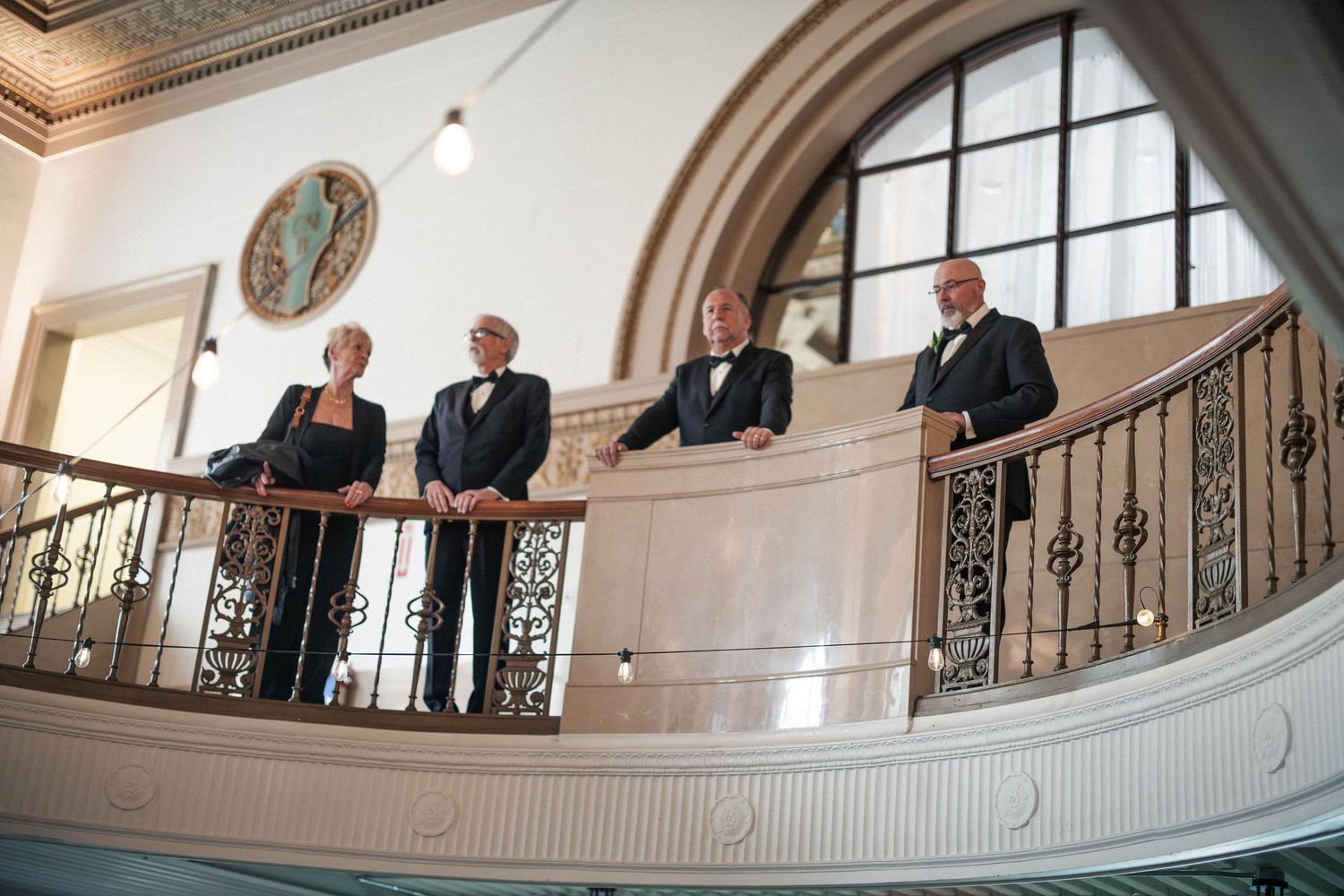 Four elegantly dressed people stand on a grand staircase balcony inside a historic wedding venue.