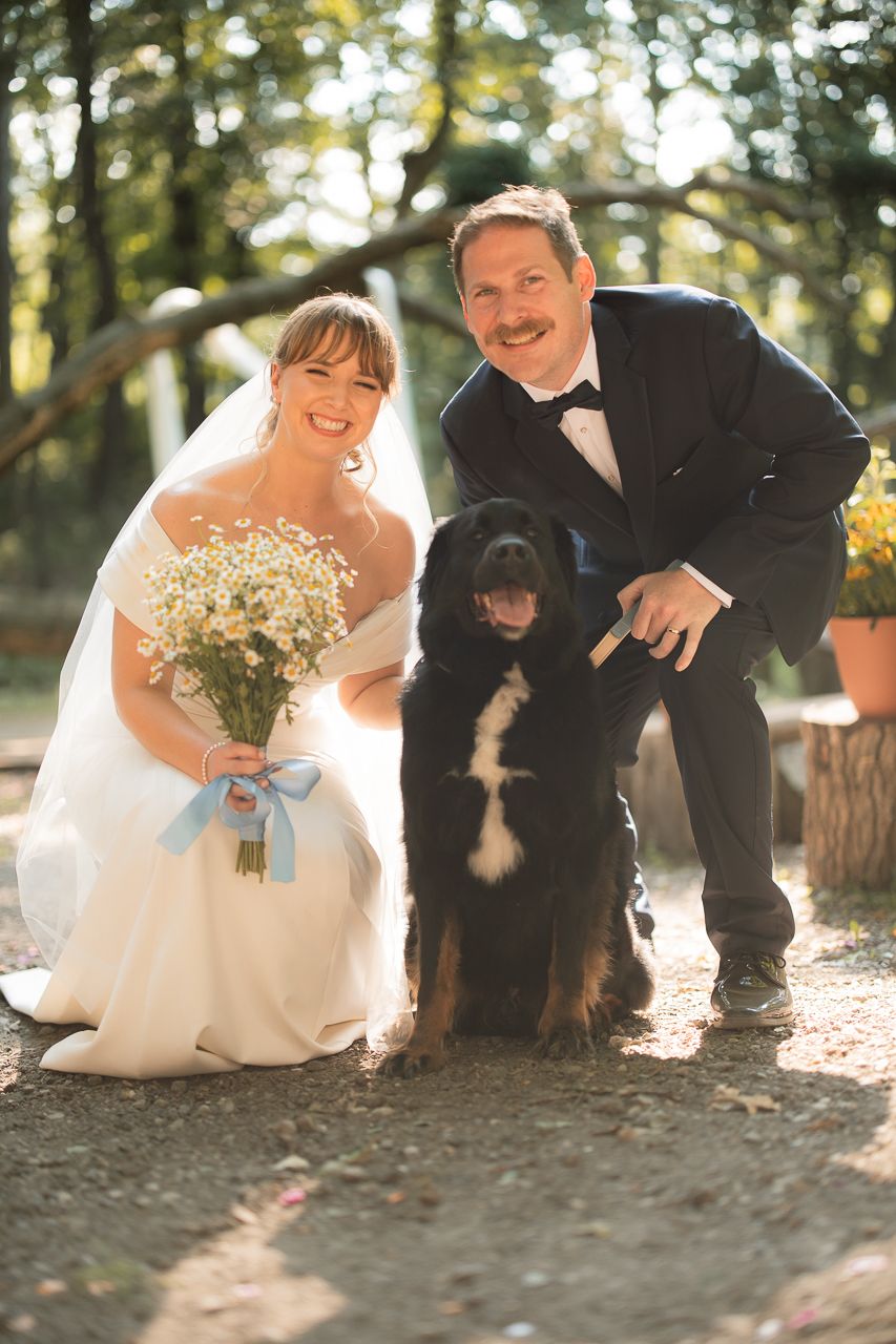 Smiling bride and groom pose with black dog at outdoor wedding, bride holding daisy bouquet.