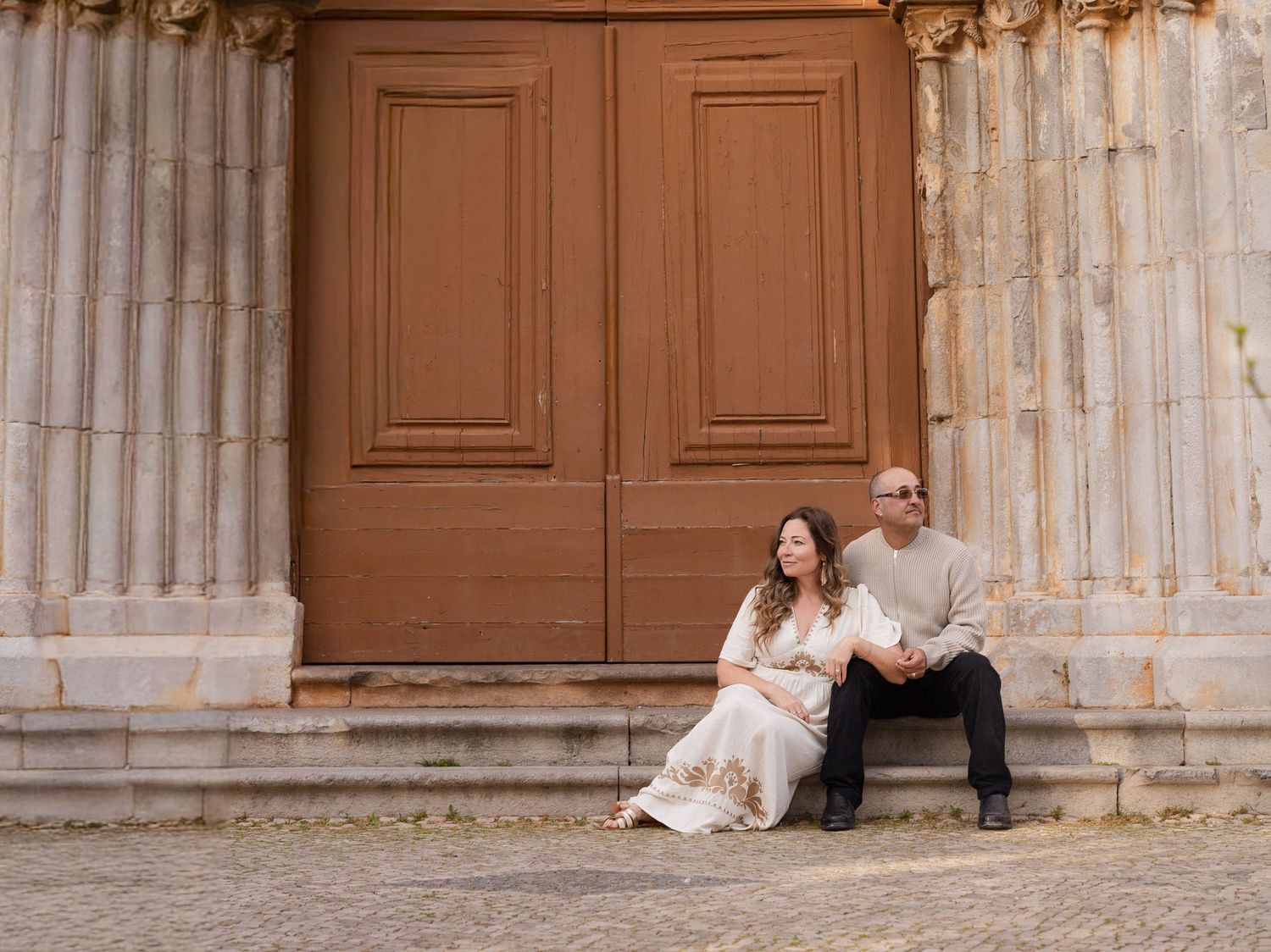 Couple sitting on stone steps in front of large wooden doors at a historic building, dressed in elegant attire.