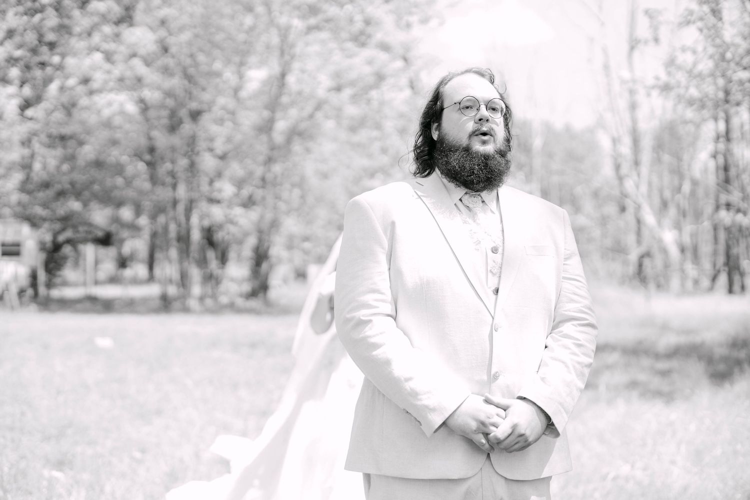 Groom in white suit standing outdoors in black and white wedding photo with trees in background.