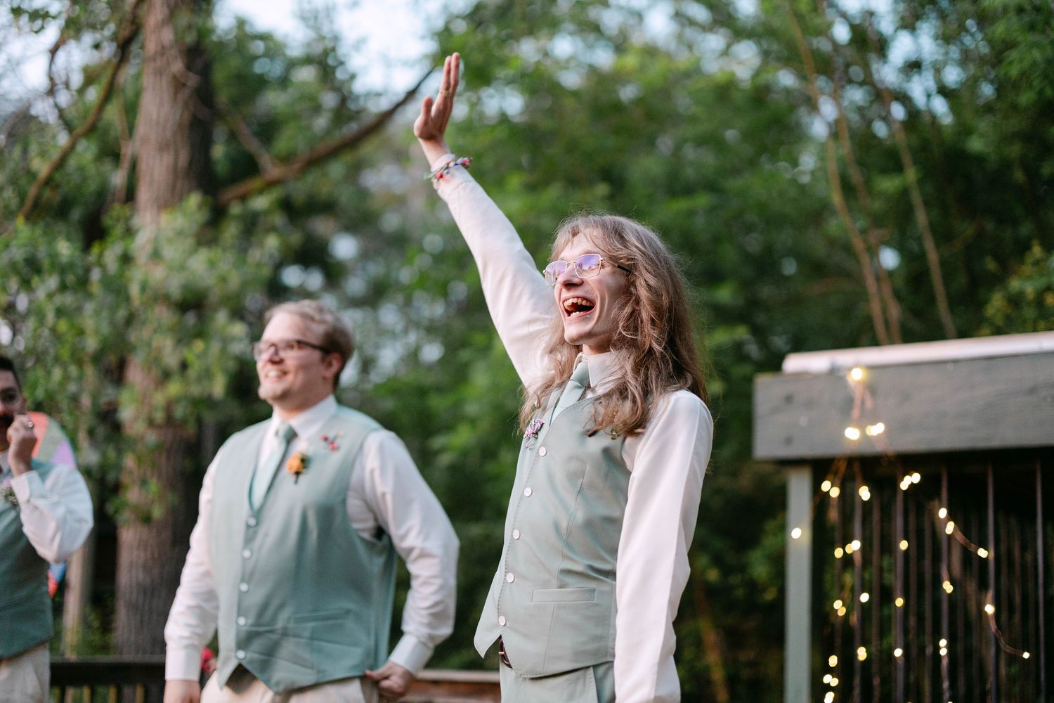 Joyful wedding guest in mint vest raises hand and laughs during an outdoor reception celebration among trees.