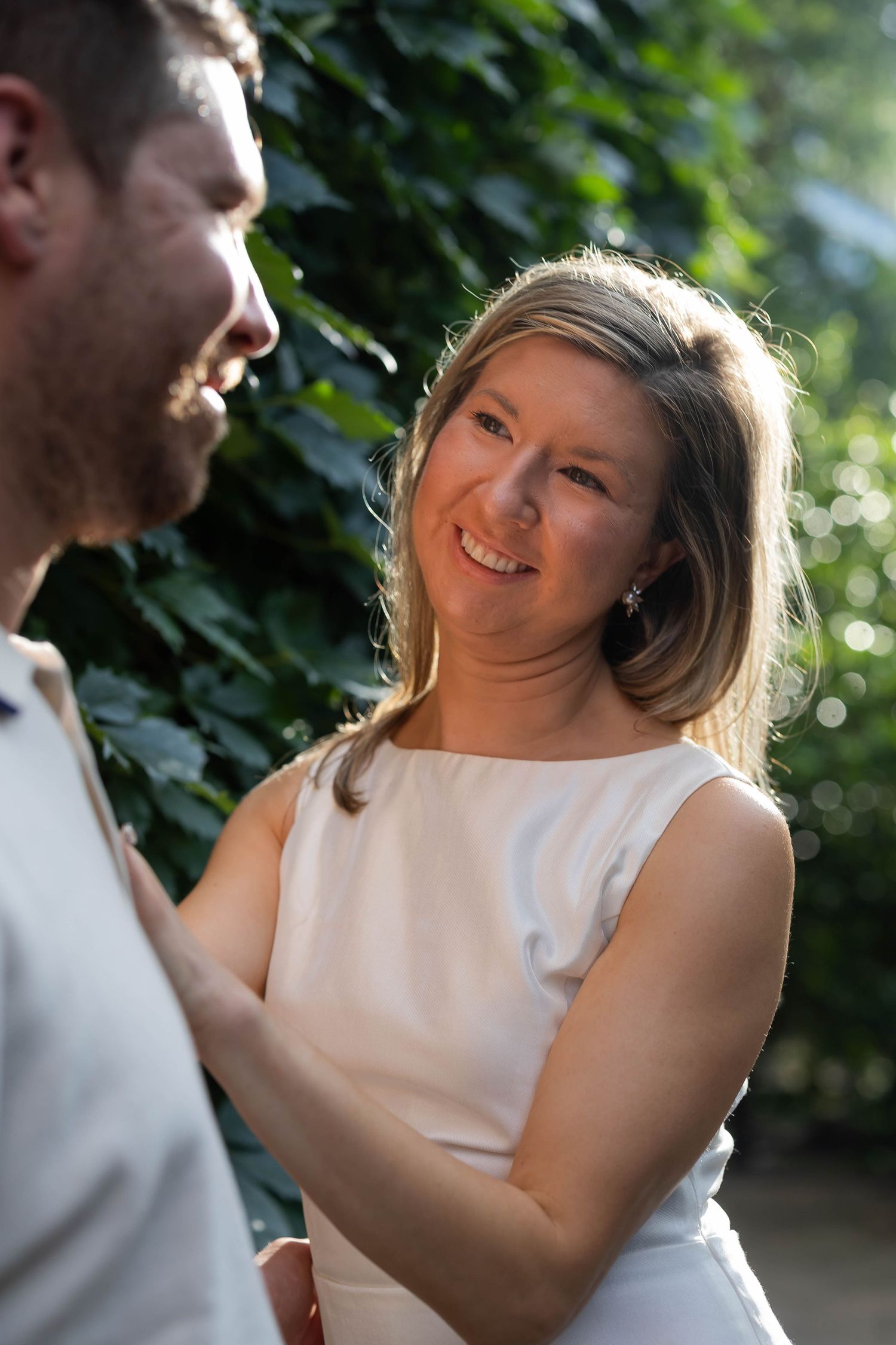 Smiling blonde woman in white dress gazes at partner outdoors near green foliage.