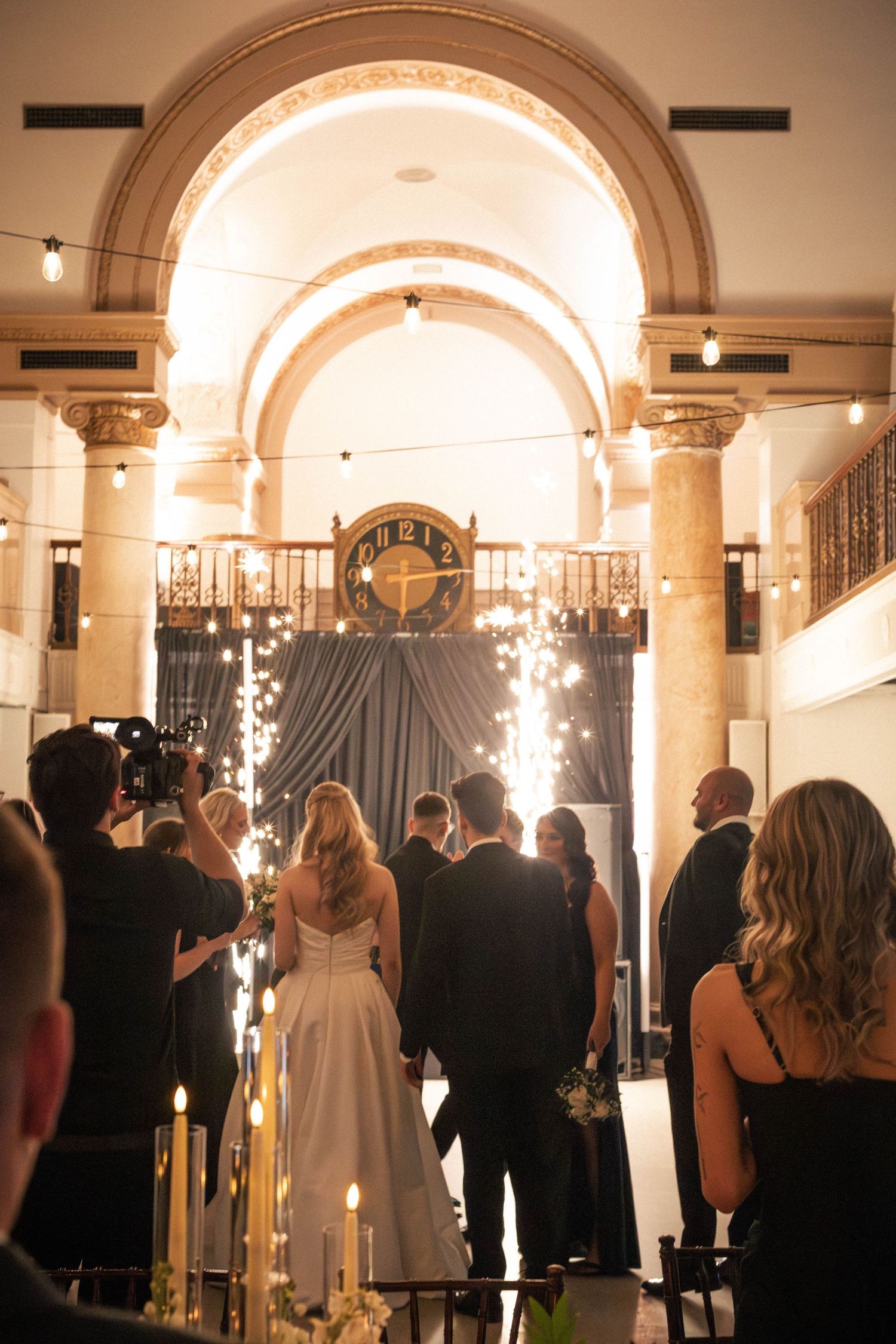 Bride and groom enter reception through grand arch with sparklers and dramatic clock backdrop at night.