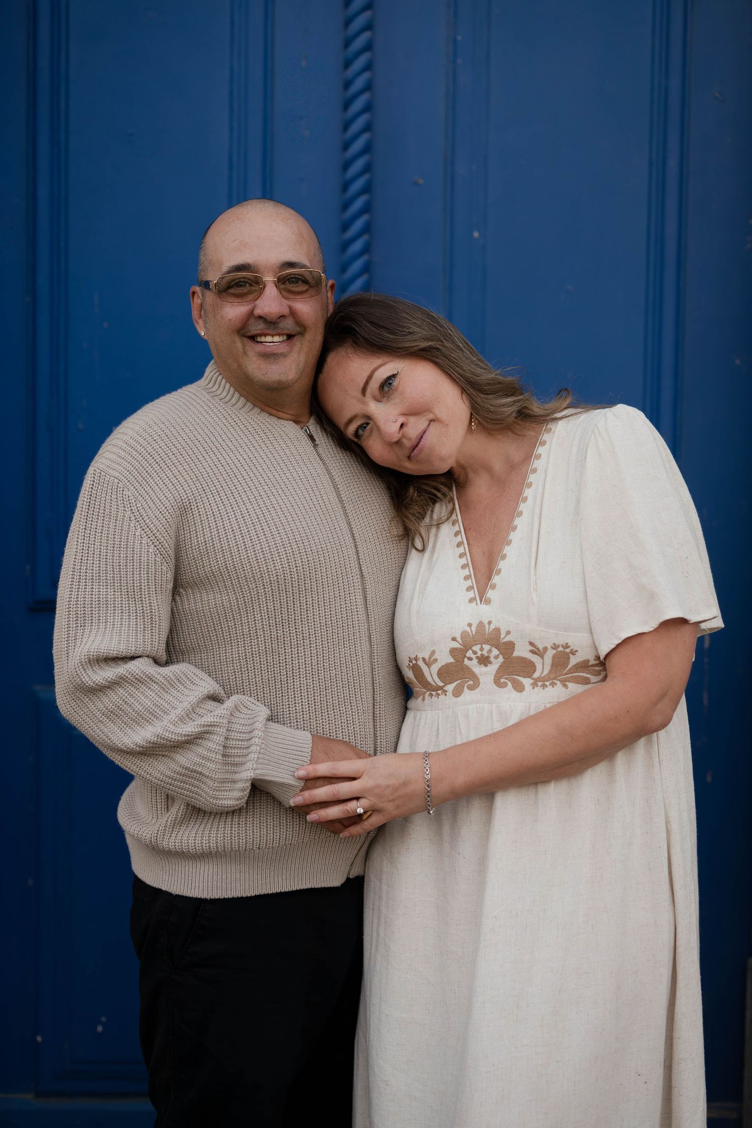 Smiling couple posing affectionately in front of a vibrant blue door, man in beige sweater, woman in white dress.