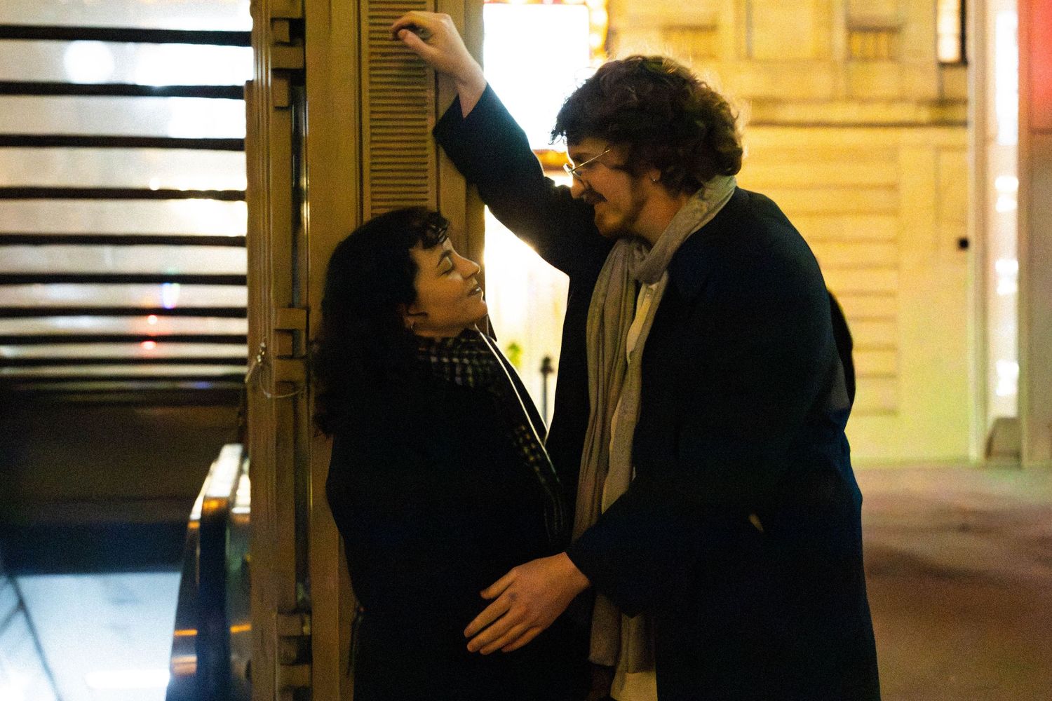 A couple sharing an intimate moment on a city street at night, the man leaning toward the woman under warm lights.
