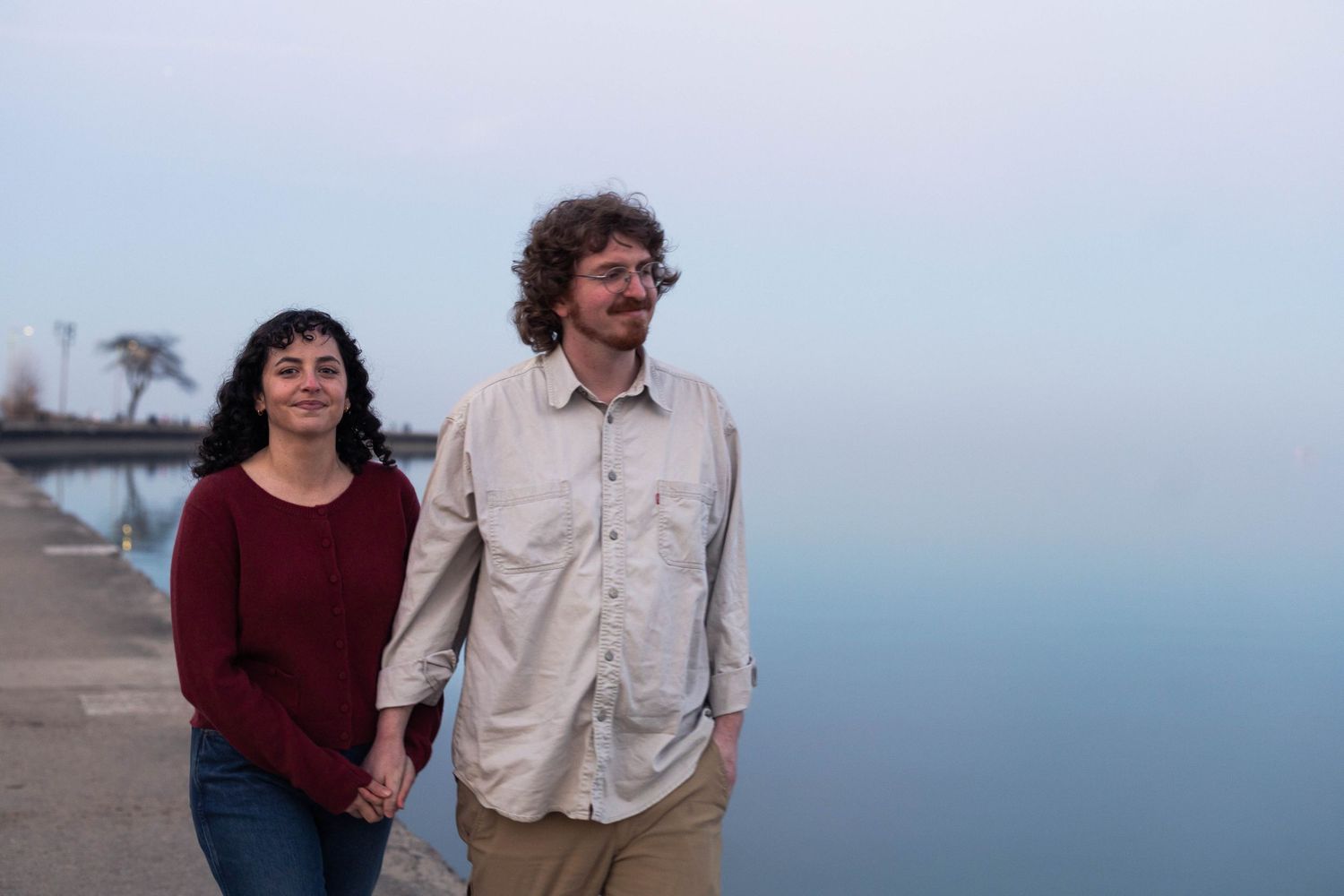 Couple holding hands walking along a waterfront promenade on a misty day near calm blue water.