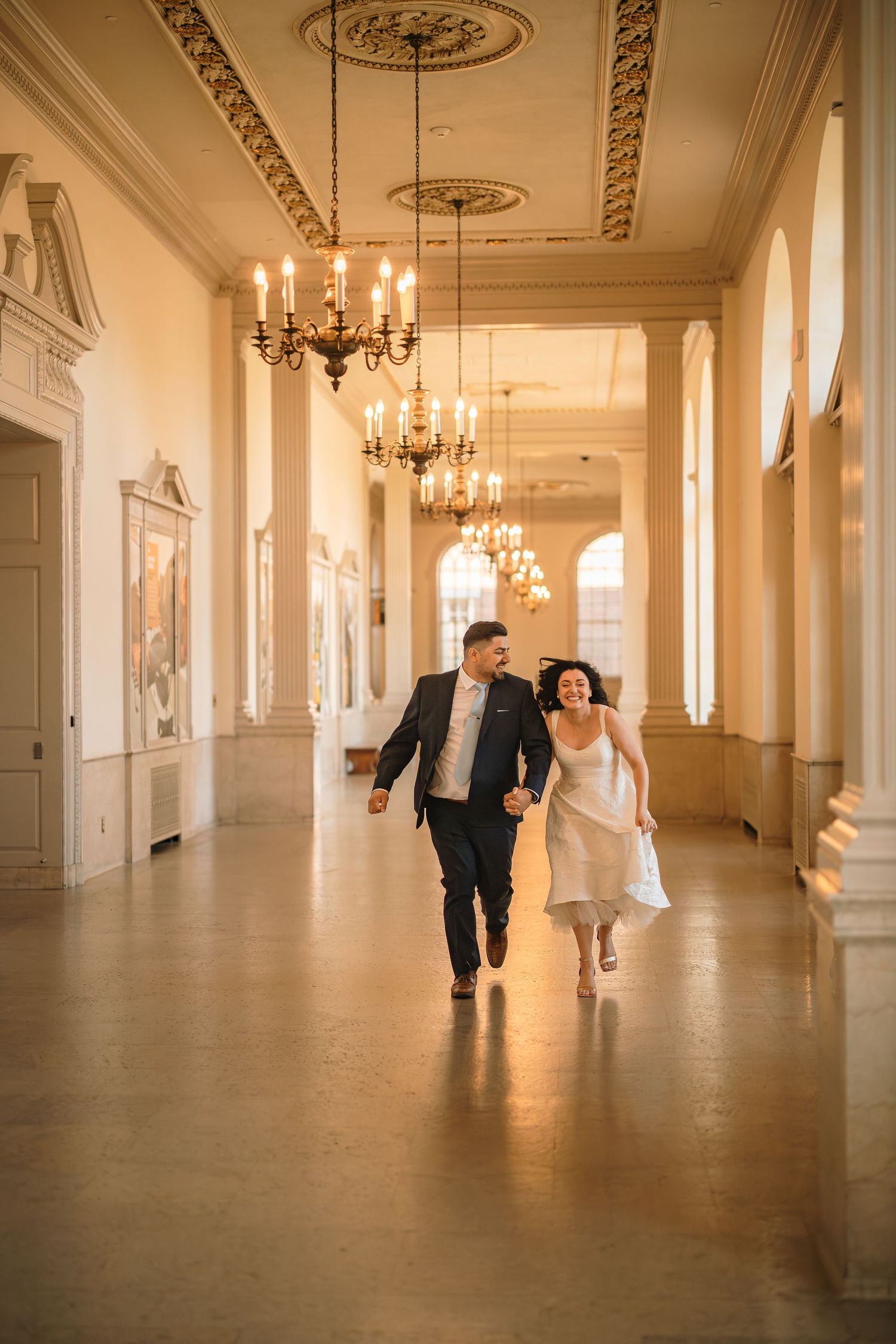 A couple walks through an elegant hallway with chandeliers and ornate moldings in a historic building.