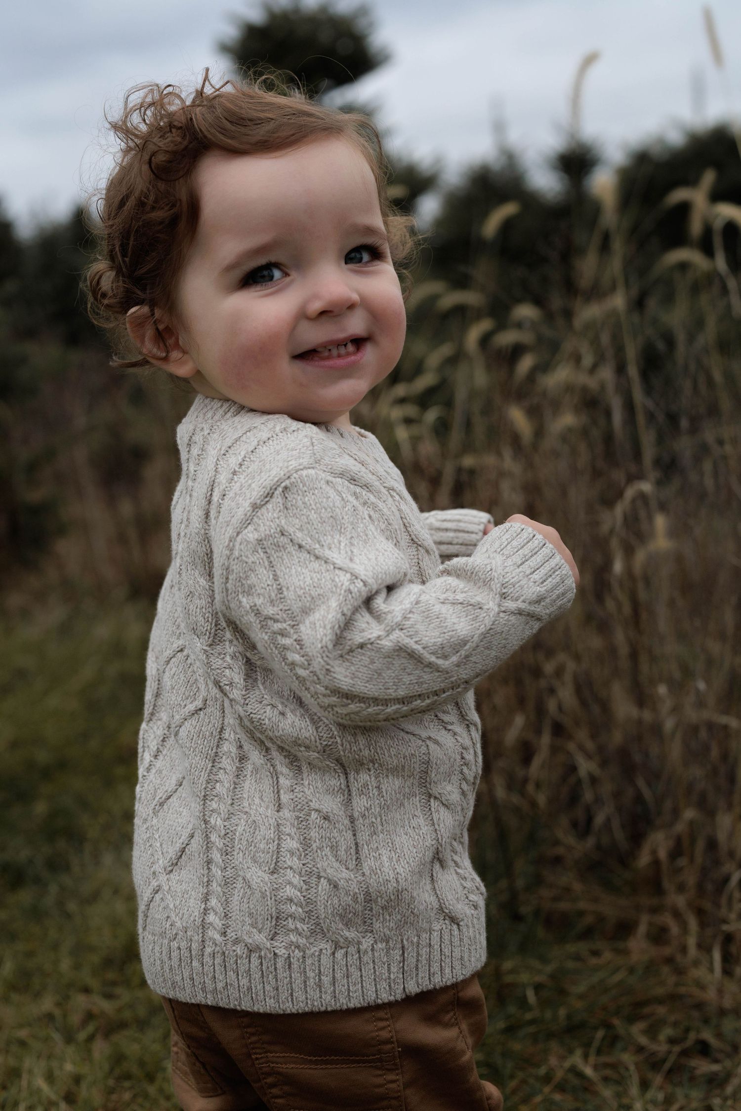 Smiling toddler in a cream cable-knit sweater standing outdoors in a field with tall grass.
