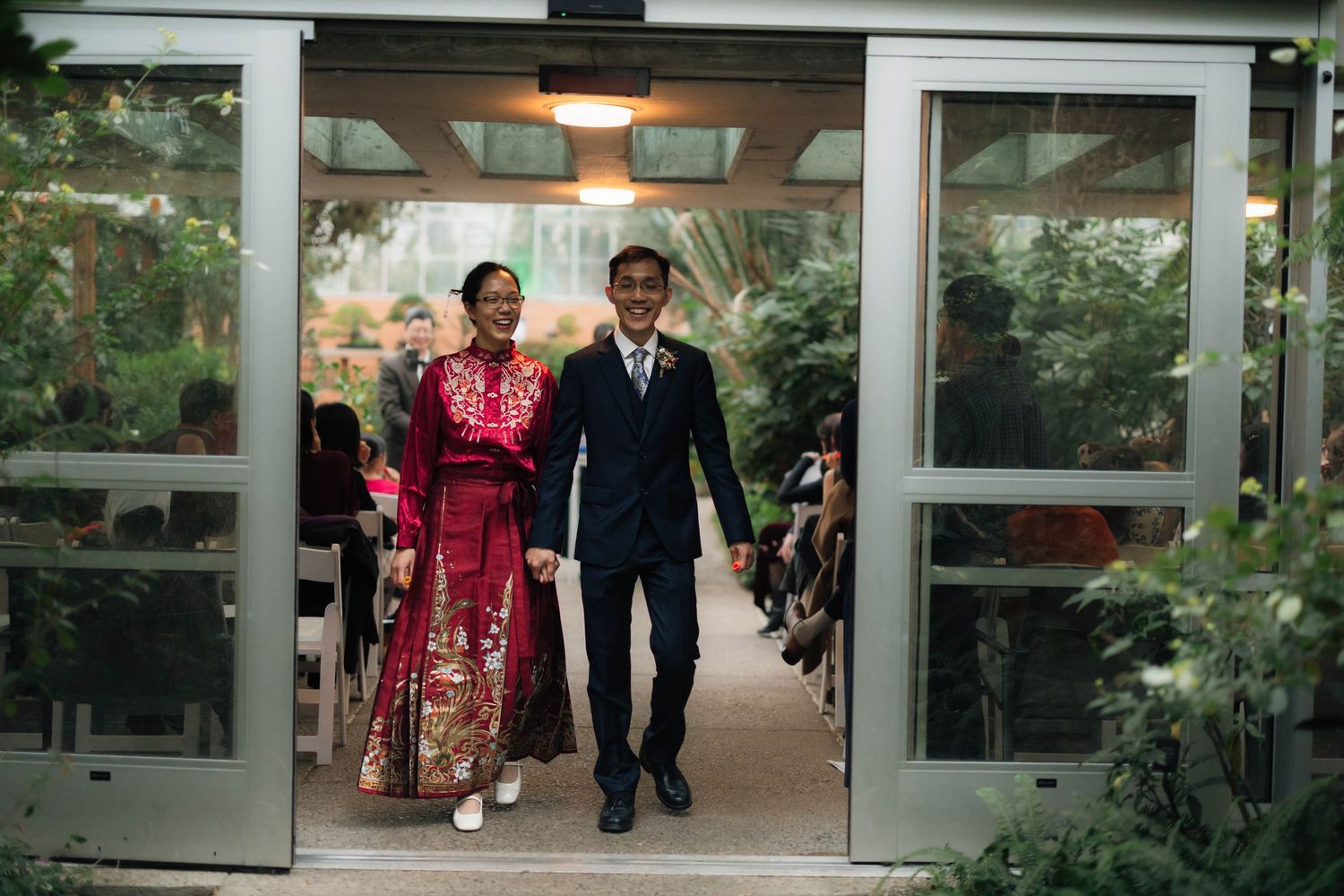 Couple in traditional Chinese wedding attire walking down aisle in greenhouse venue.