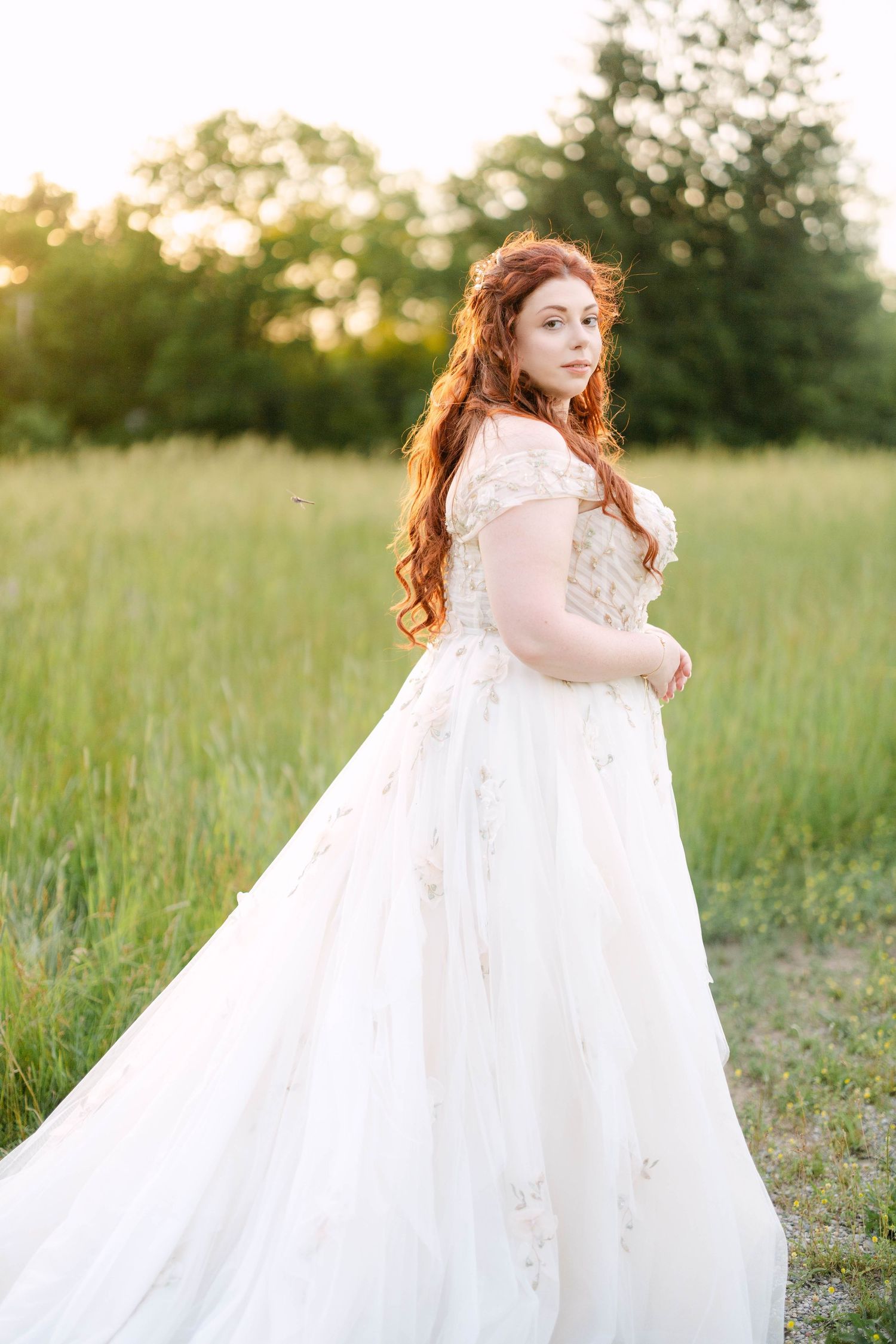 Red-haired bride in off-shoulder white ballgown standing in sunlit green meadow at golden hour.