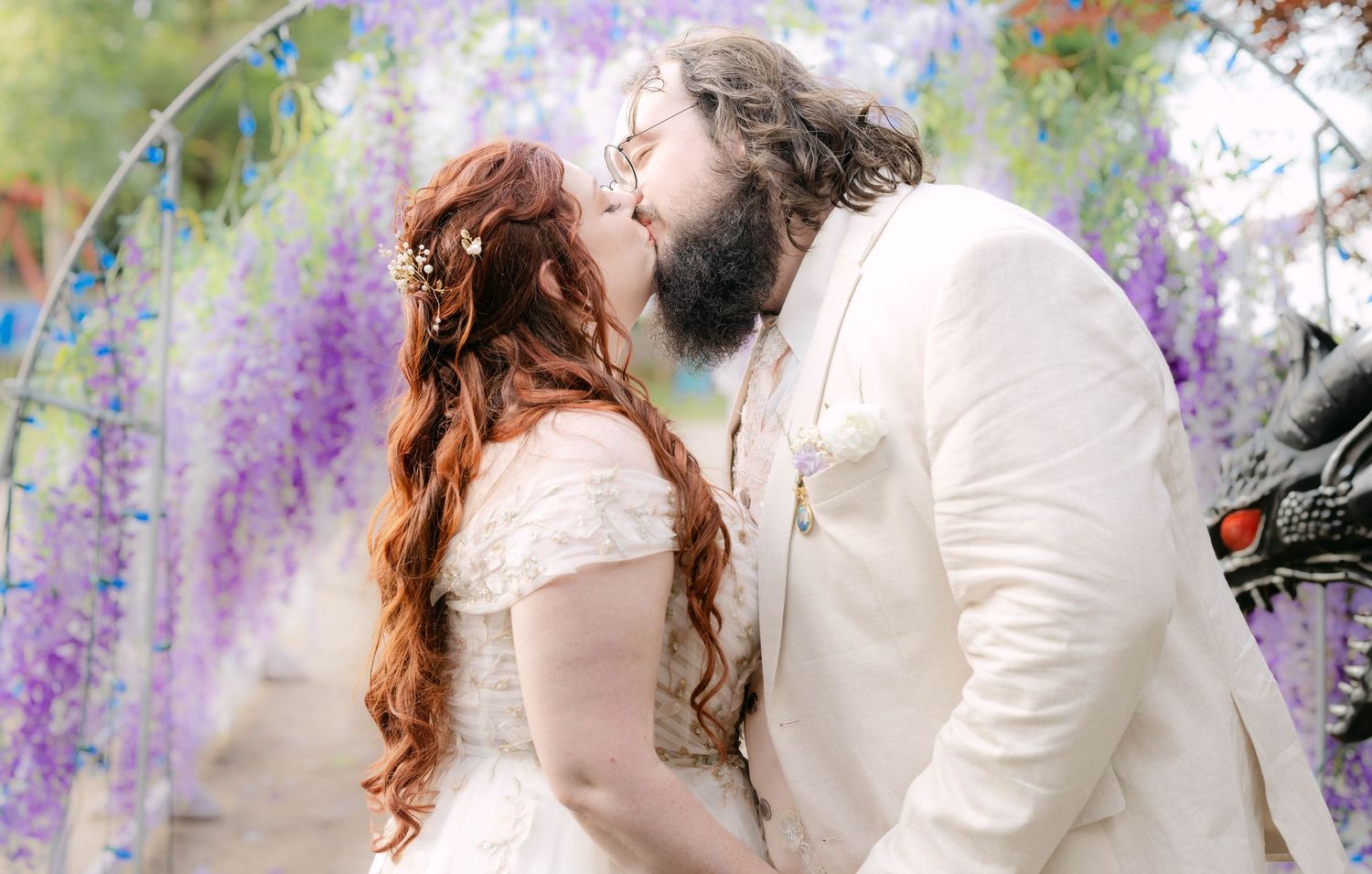 A couple shares a romantic kiss under a stunning wisteria arch in full purple bloom on their wedding day.