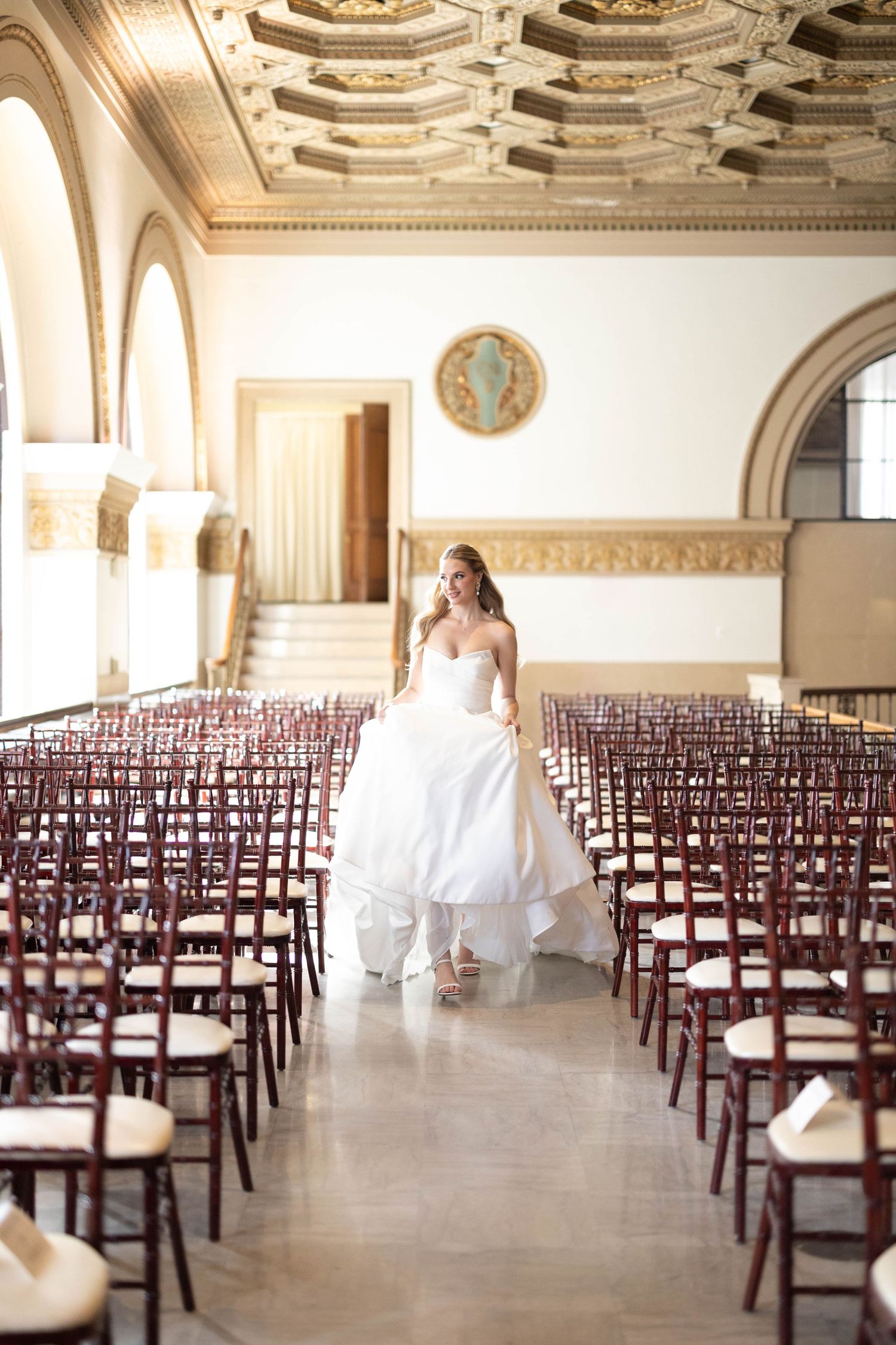 Bride in white ball gown walks down aisle of grand ornate venue with rows of dark wooden chairs.