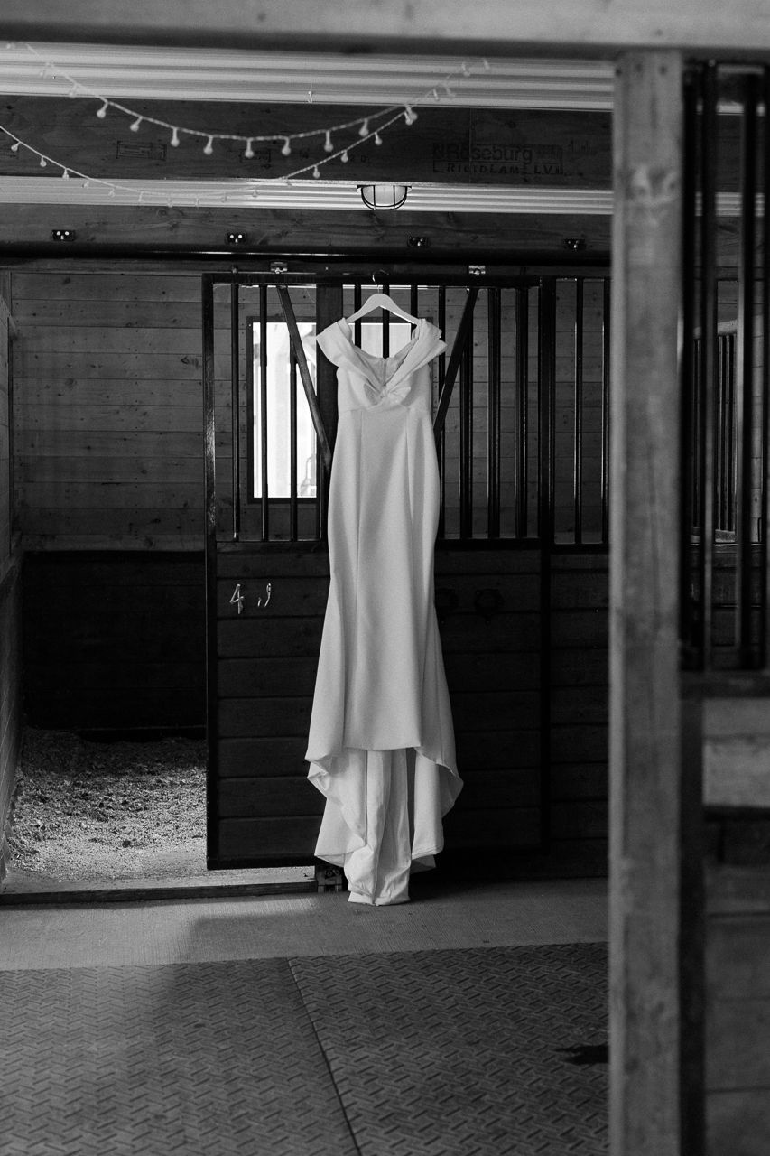 Black and white photo of an elegant wedding gown hanging in a rustic barn stable doorway.