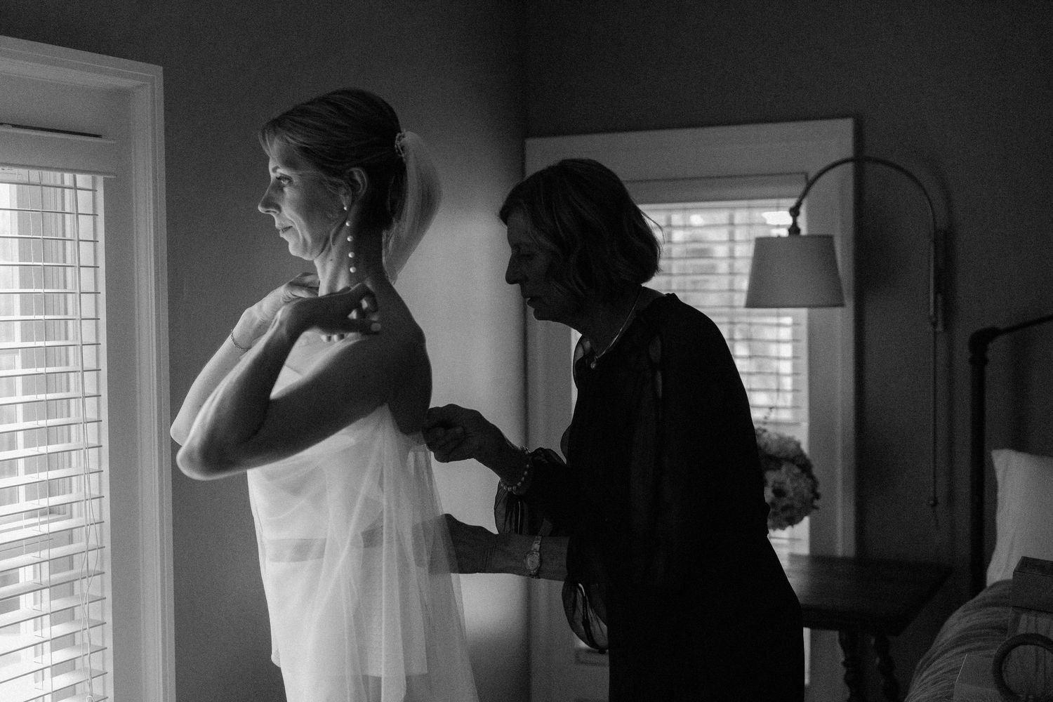 People in formal attire gather near windows in a black and white photograph during a social event.