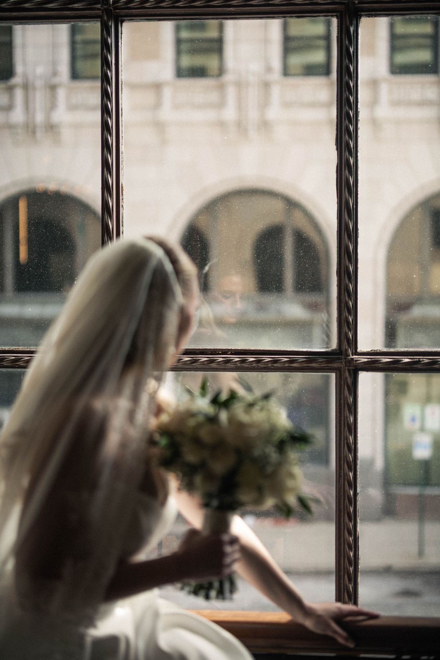 Bride in white gown and veil holds bouquet while gazing out a large vintage window on her wedding day.