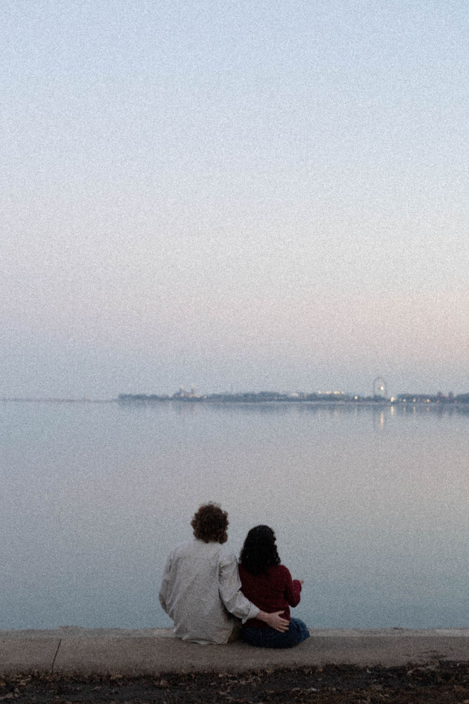 Couple sitting together on a waterfront ledge at dusk, gazing at a calm lake with a distant city skyline.