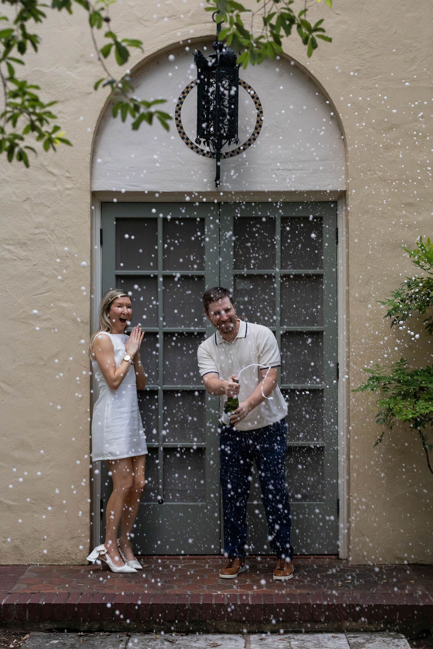 Couple celebrating with champagne spray in front of an arched door, woman in white dress, man in casual attire.