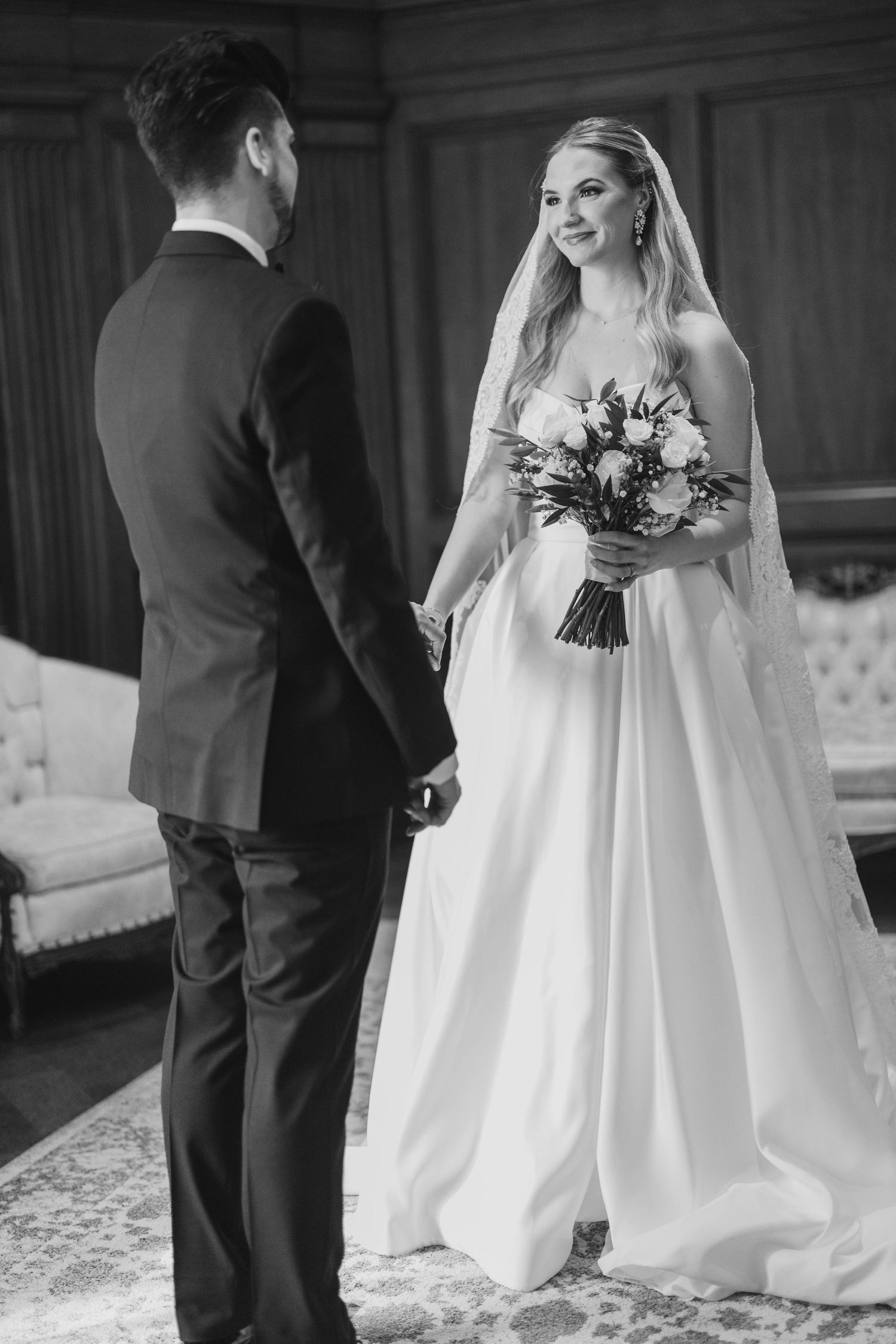 Black and white photo of a smiling bride holding bouquet facing groom in suit during wedding ceremony.