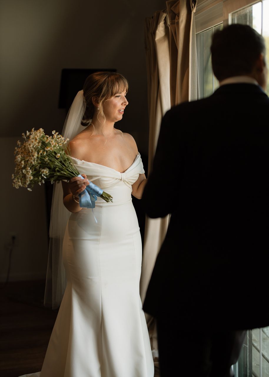 Elegant bride in off-shoulder white gown holds bouquet during emotional first look moment.