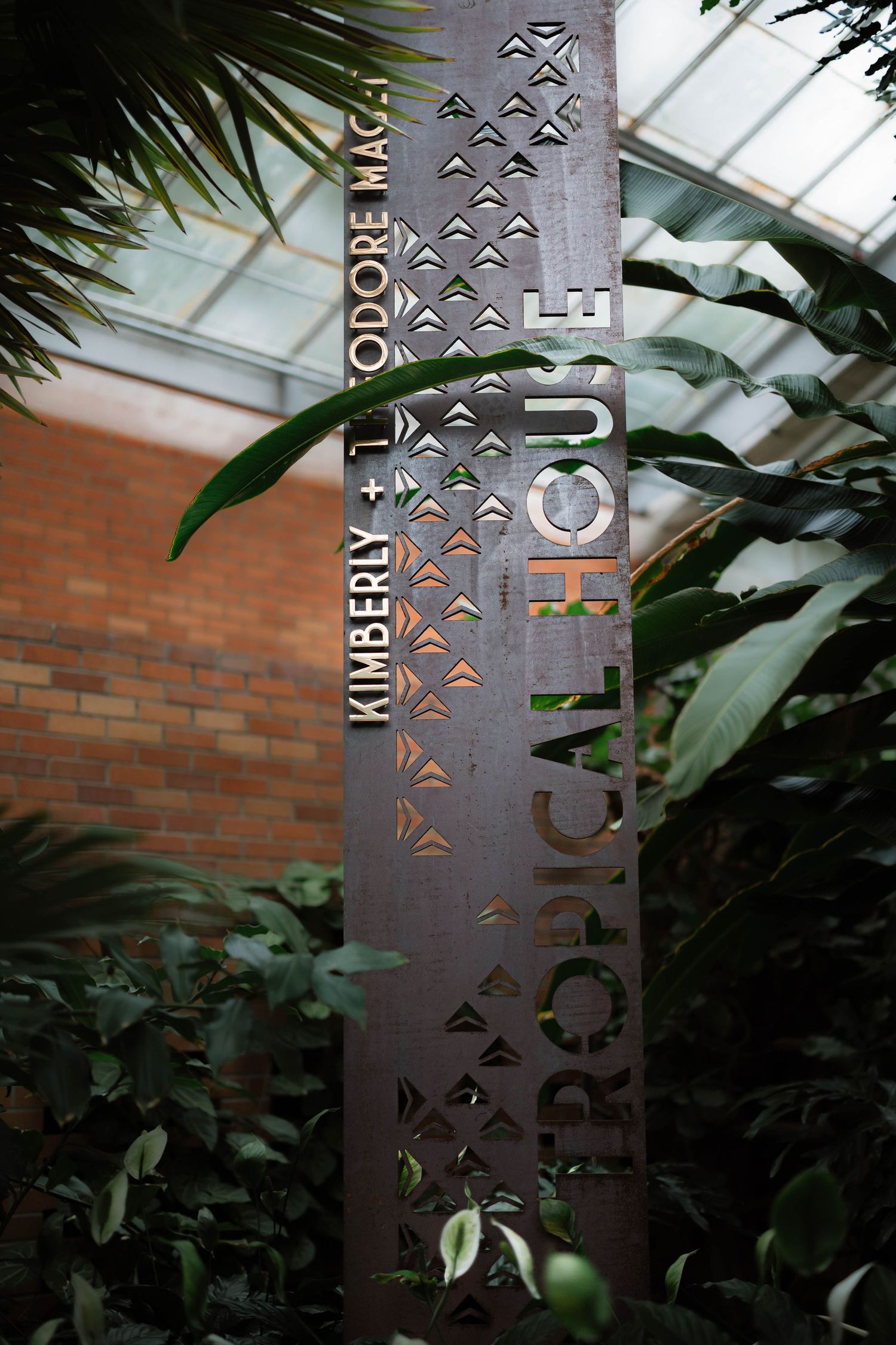Metal sign for Tropical House dedicated to Kimberly and Theodore Mack surrounded by tropical plants.