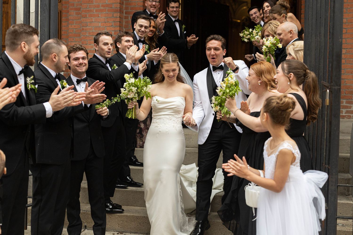 Bride and groom exit church surrounded by cheering wedding party in black attire, holding white floral bouquets.
