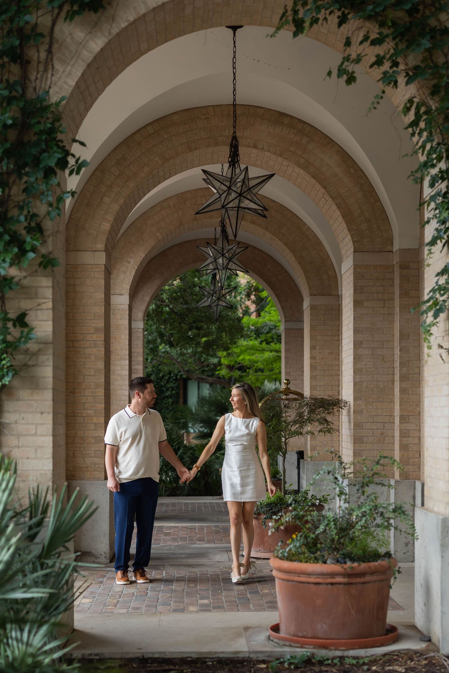 Couple holding hands under elegant brick arches with star pendant light and lush greenery, dressed in white outfits.