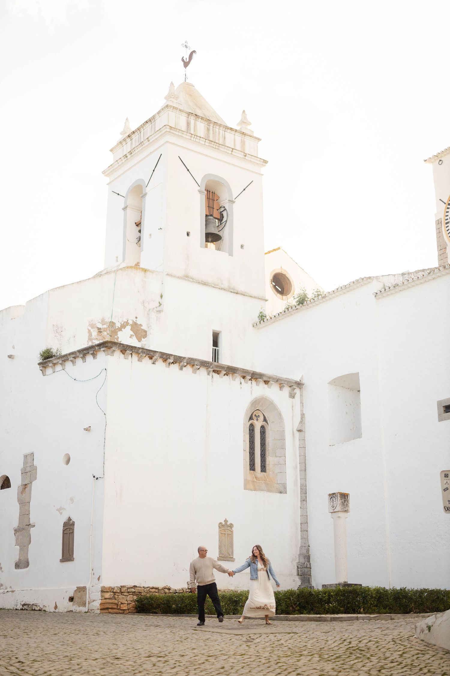 Couple holding hands walking in front of a white historic church with bell tower on cobblestone square.