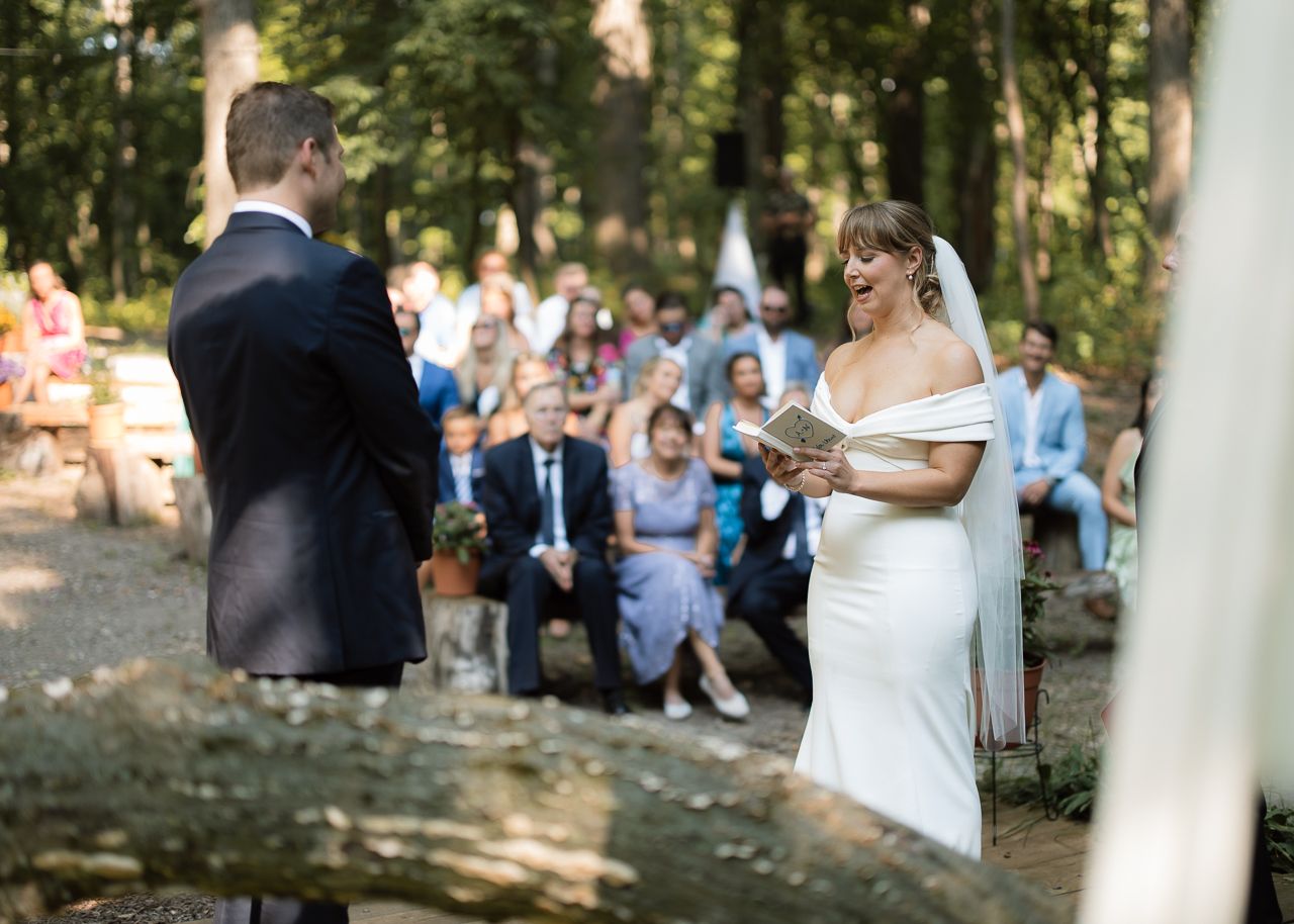 Bride in white off-shoulder gown reads vows to groom during outdoor forest wedding ceremony with guests seated behind.