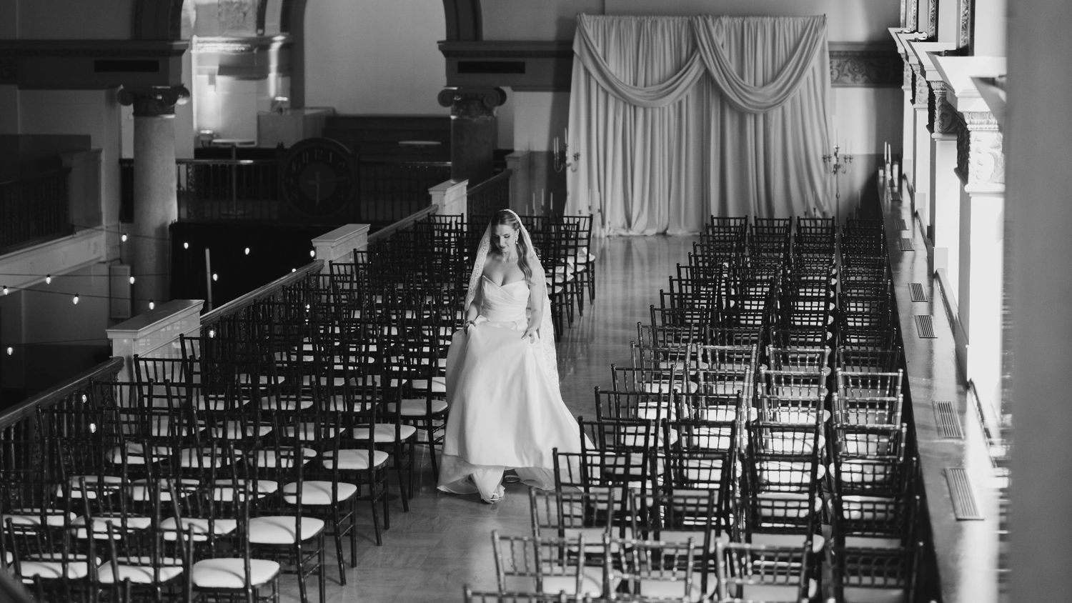 Black and white photo of a bride in a white gown and veil standing in an elegant empty venue with rows of chairs.