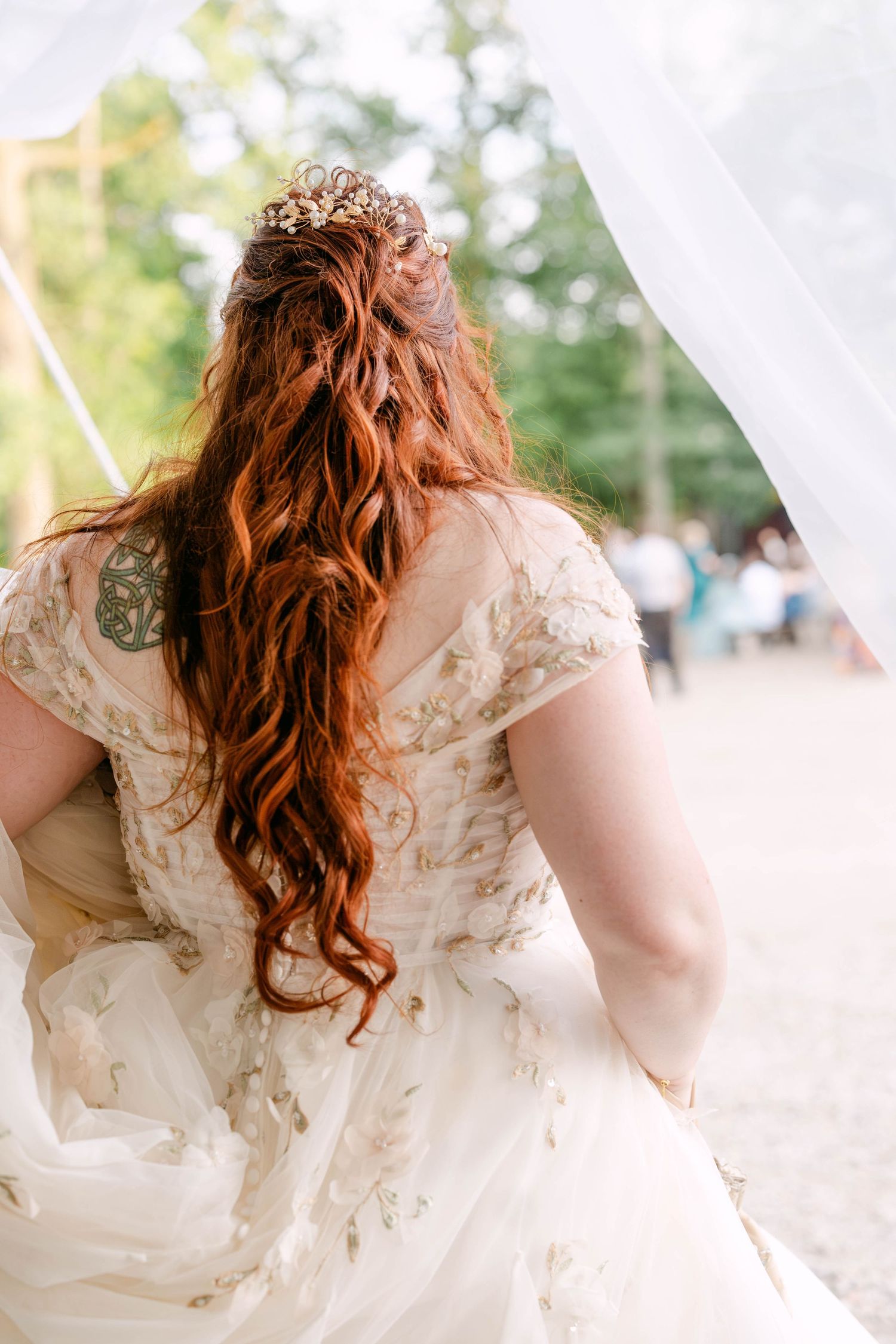 Bride with long red hair, gold crown, and floral wedding dress viewed from behind at outdoor ceremony.
