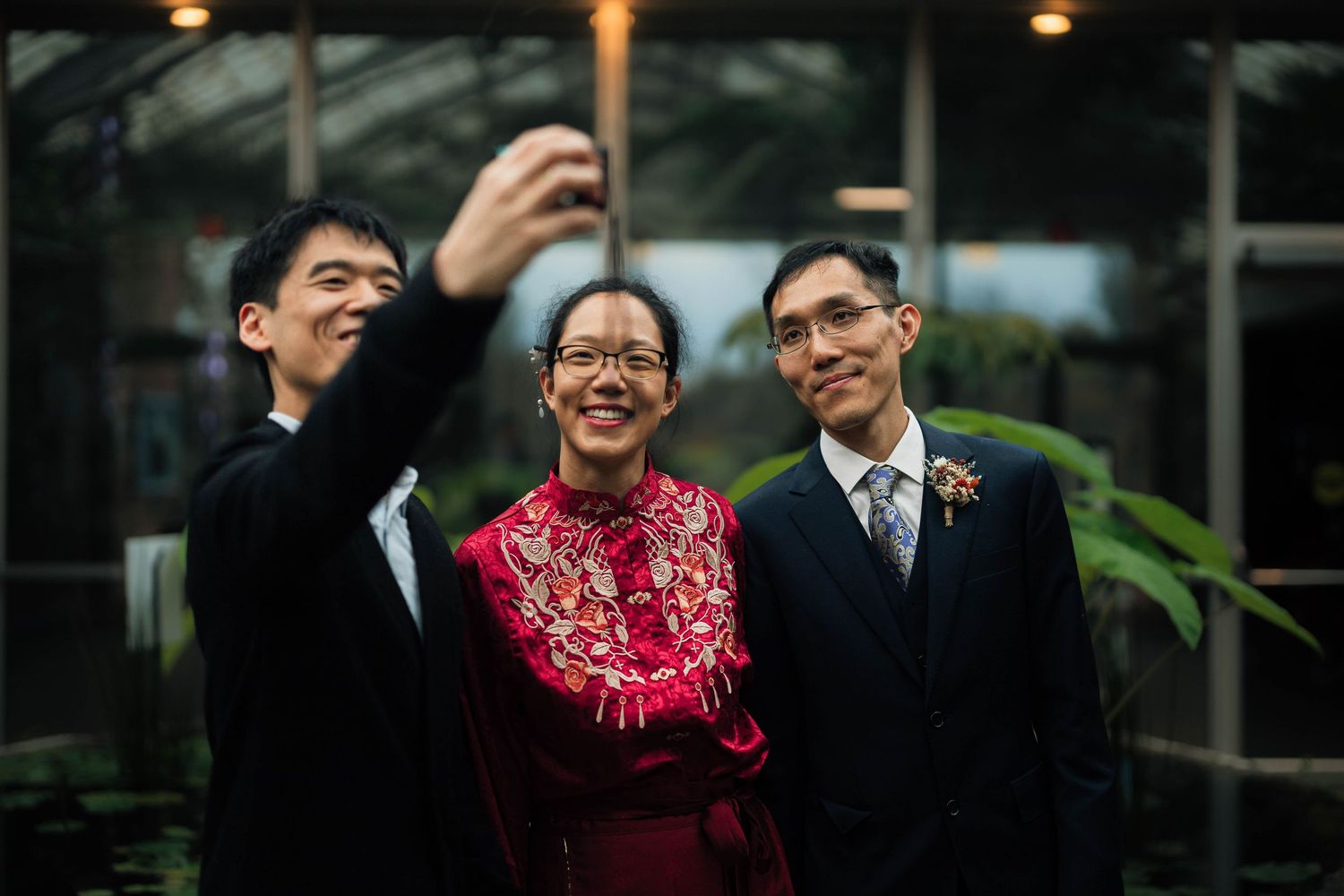 Three people take a joyful selfie outdoors, with woman in red traditional dress flanked by two men in suits.