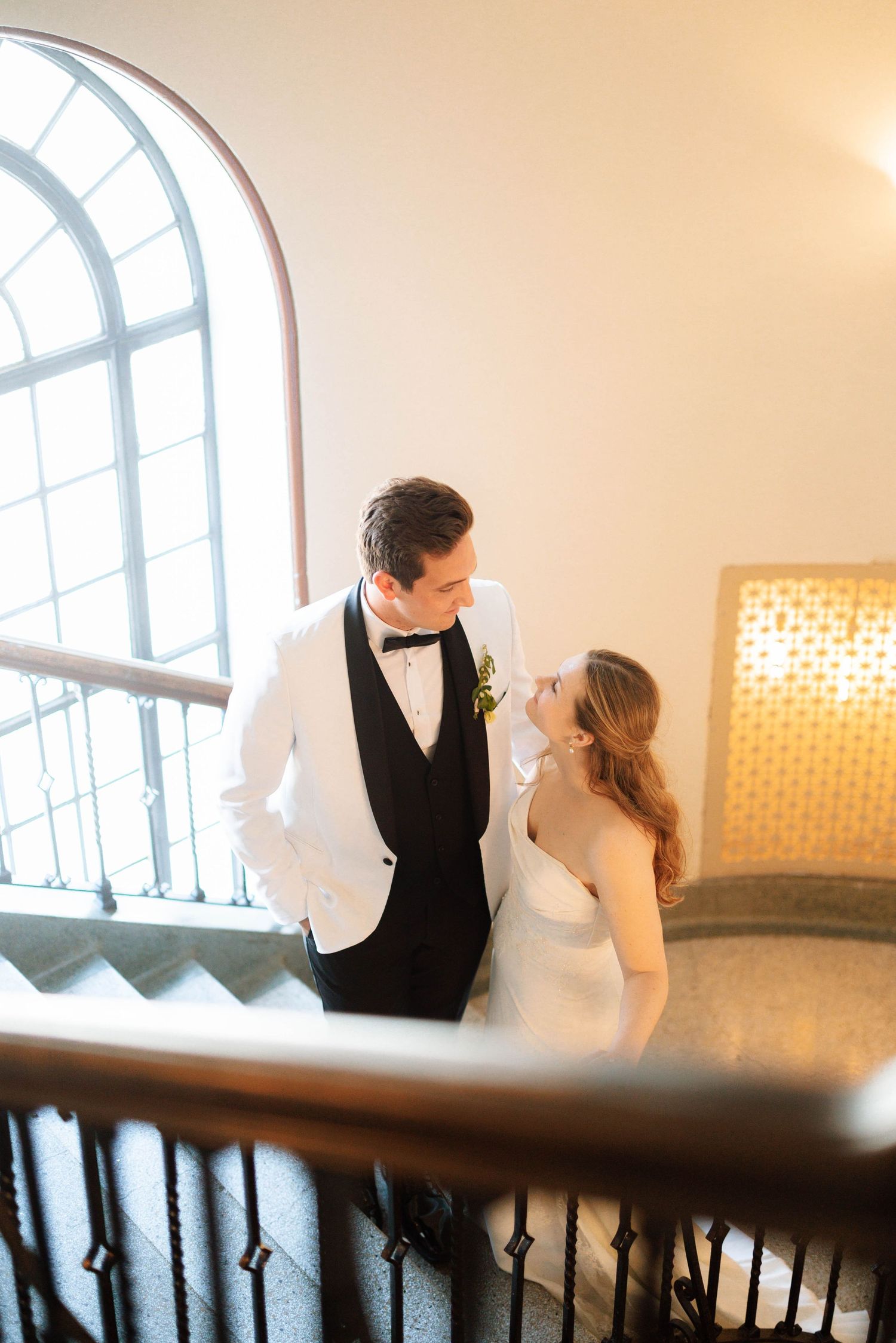 Bride and groom share a romantic moment on a grand staircase, groom in white tuxedo jacket, bride in elegant white gown.