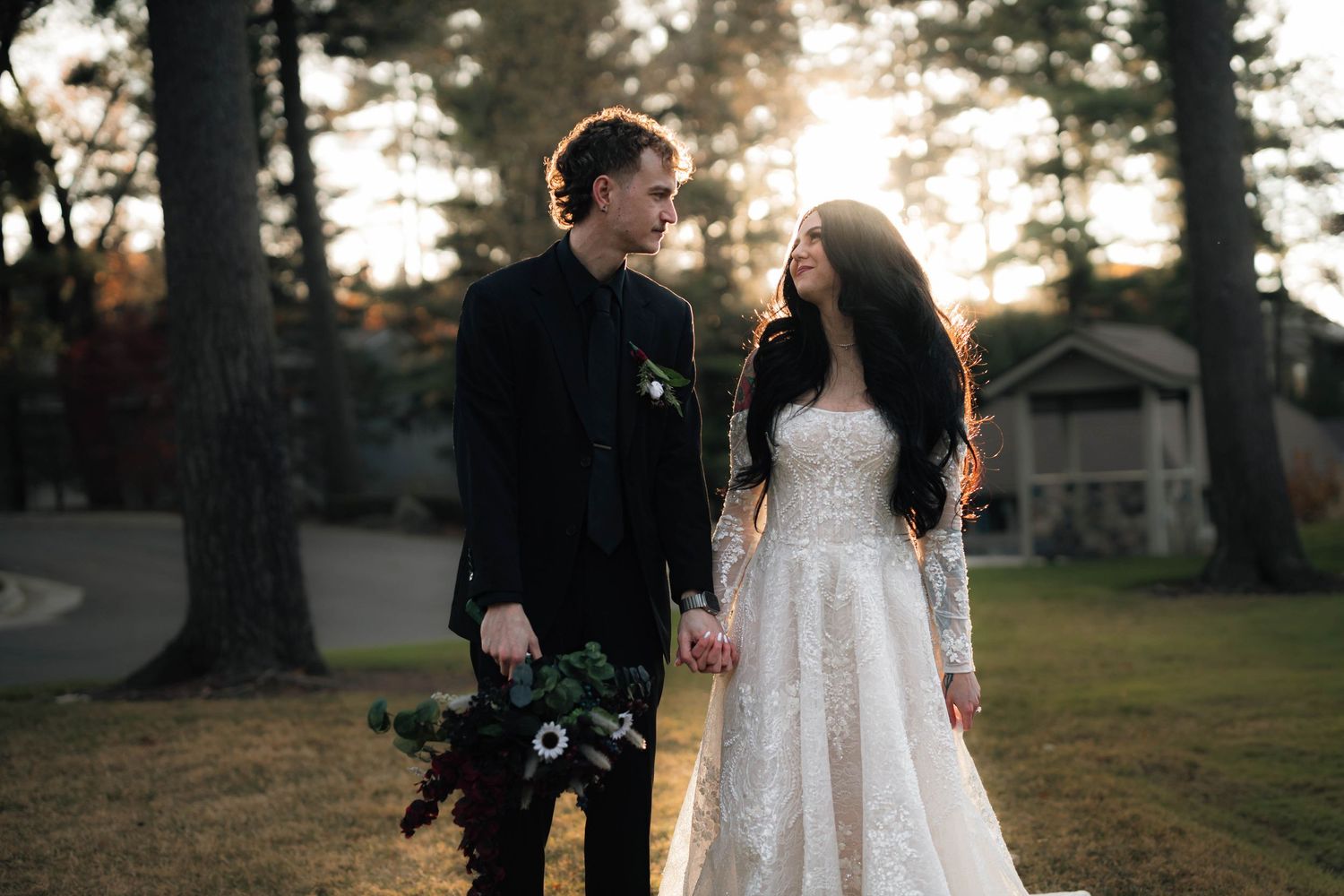 A bride in a lace gown and groom in black suit hold hands outdoors at golden hour among tall trees.