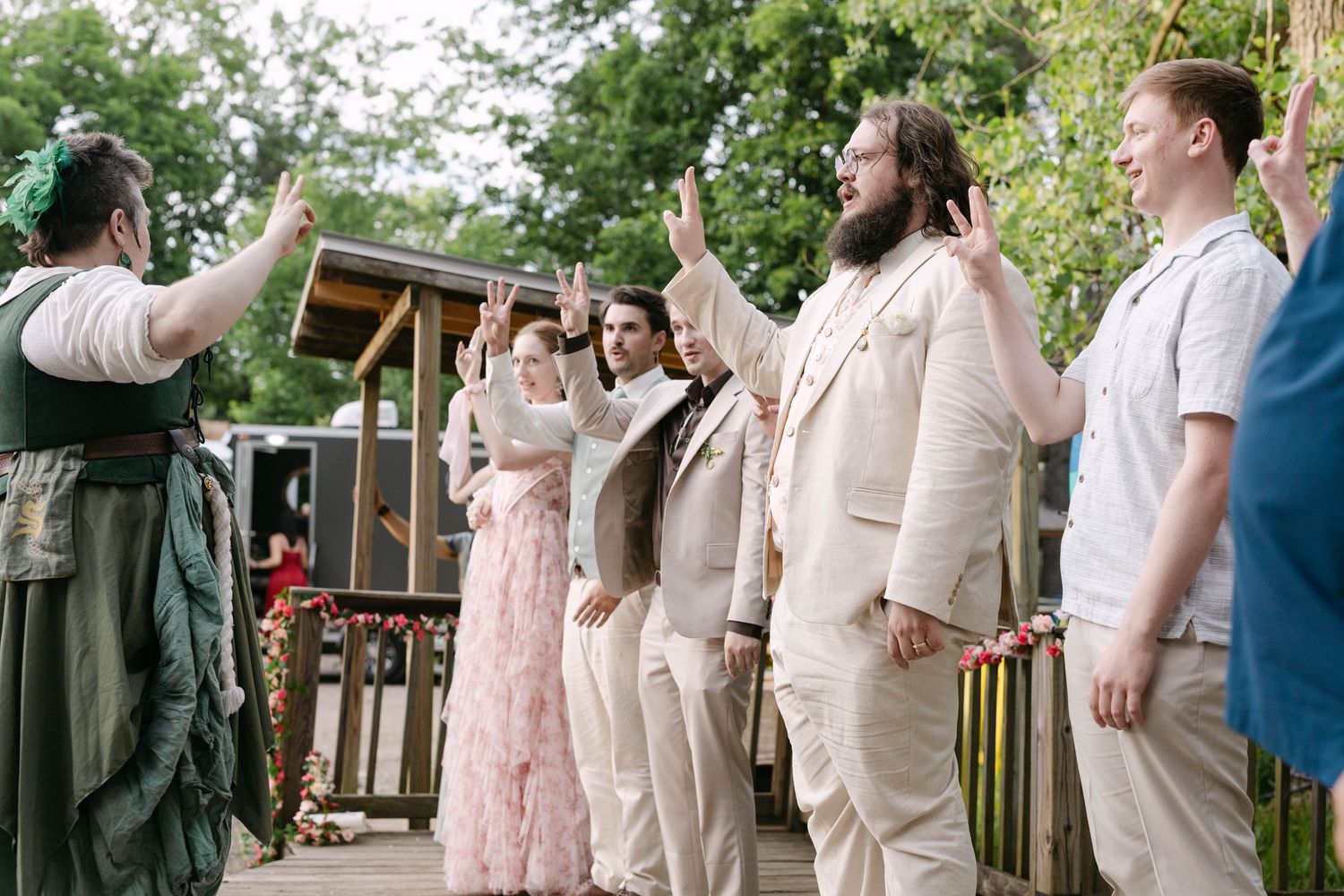 Wedding party raises hands in oath during outdoor ceremony, wearing beige suits and pink dress among lush greenery.