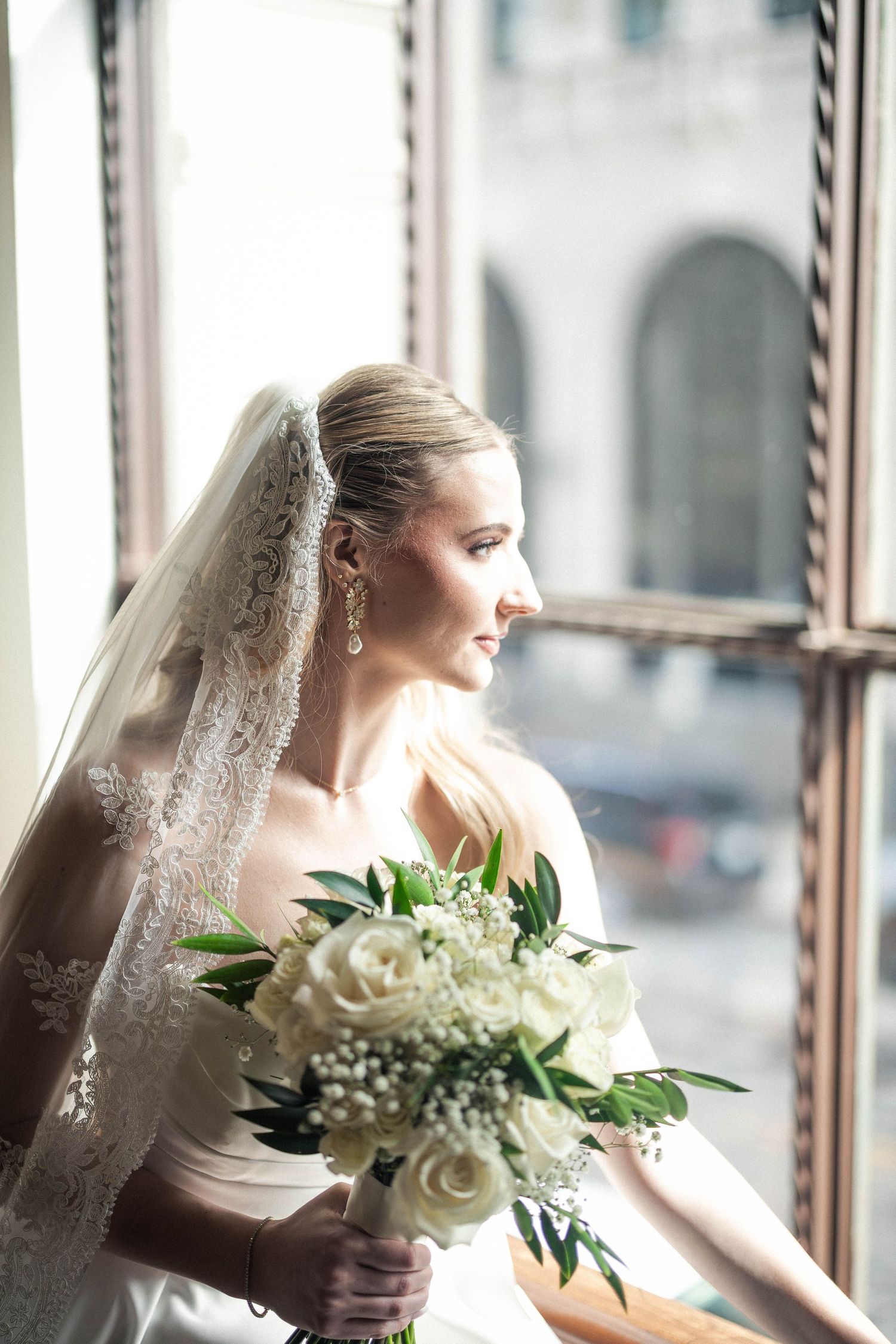 Elegant bride wearing lace veil holds white rose bouquet while gazing out window on wedding day.