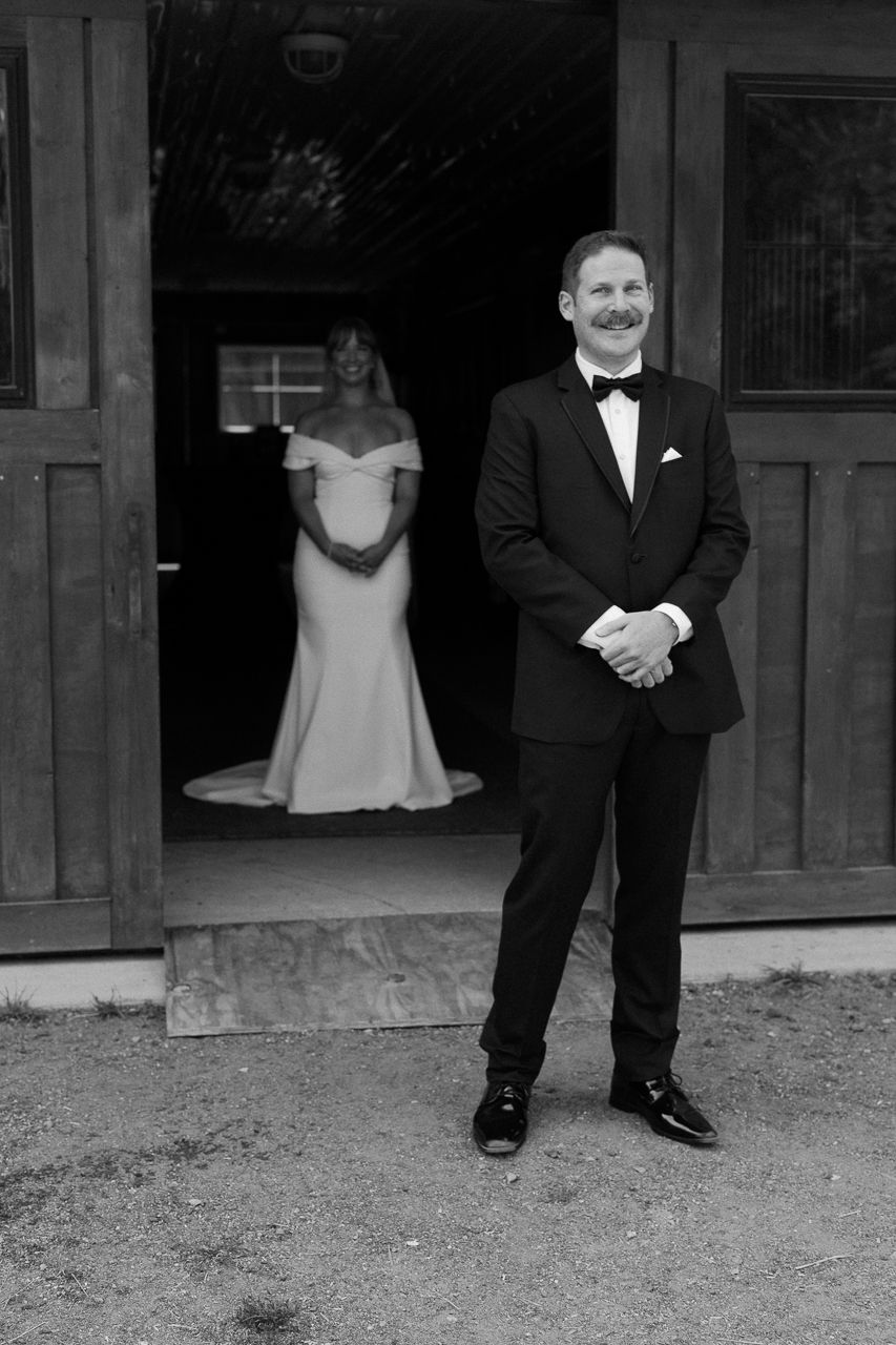 Black and white photo of a groom in a tuxedo smiling as his bride stands in a doorway behind him.